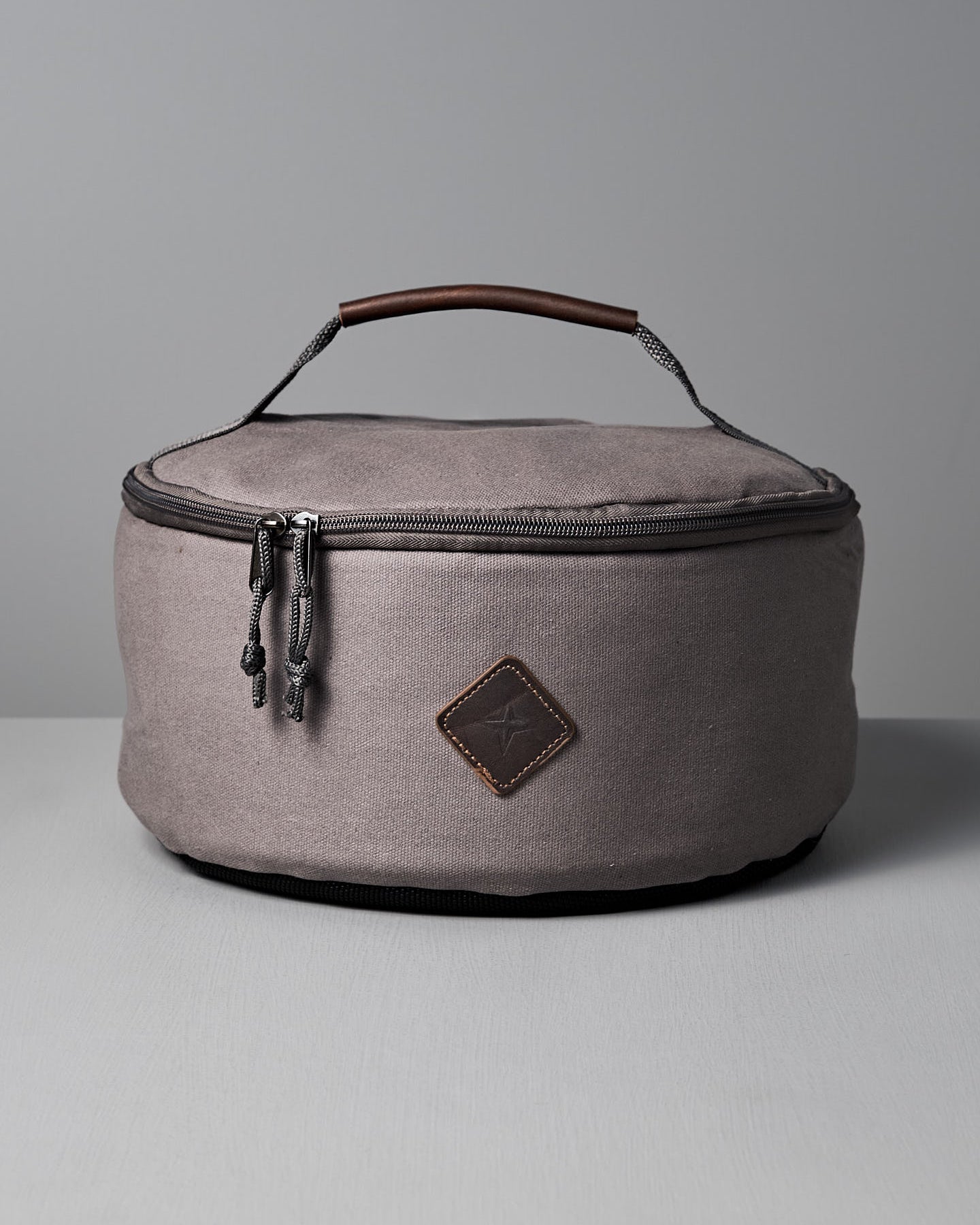 The Barebones Padded Utility Storage Bag is round and gray, made of durable cotton canvas with a zipper, brown accent patch, and top handle. It’s shown on a light gray surface against a plain background.