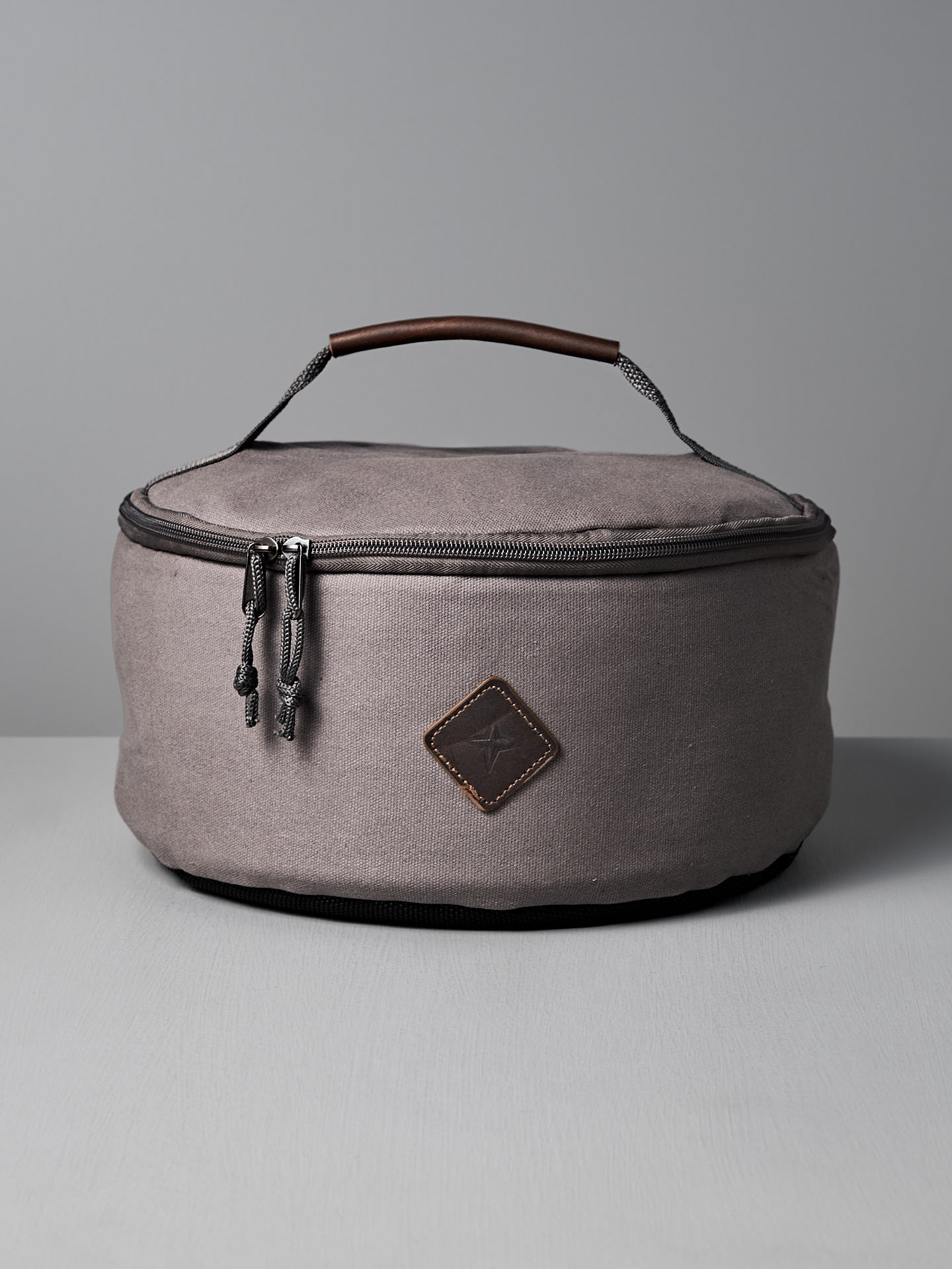 The Barebones Padded Utility Storage Bag is round and gray, made of durable cotton canvas with a zipper, brown accent patch, and top handle. It’s shown on a light gray surface against a plain background.