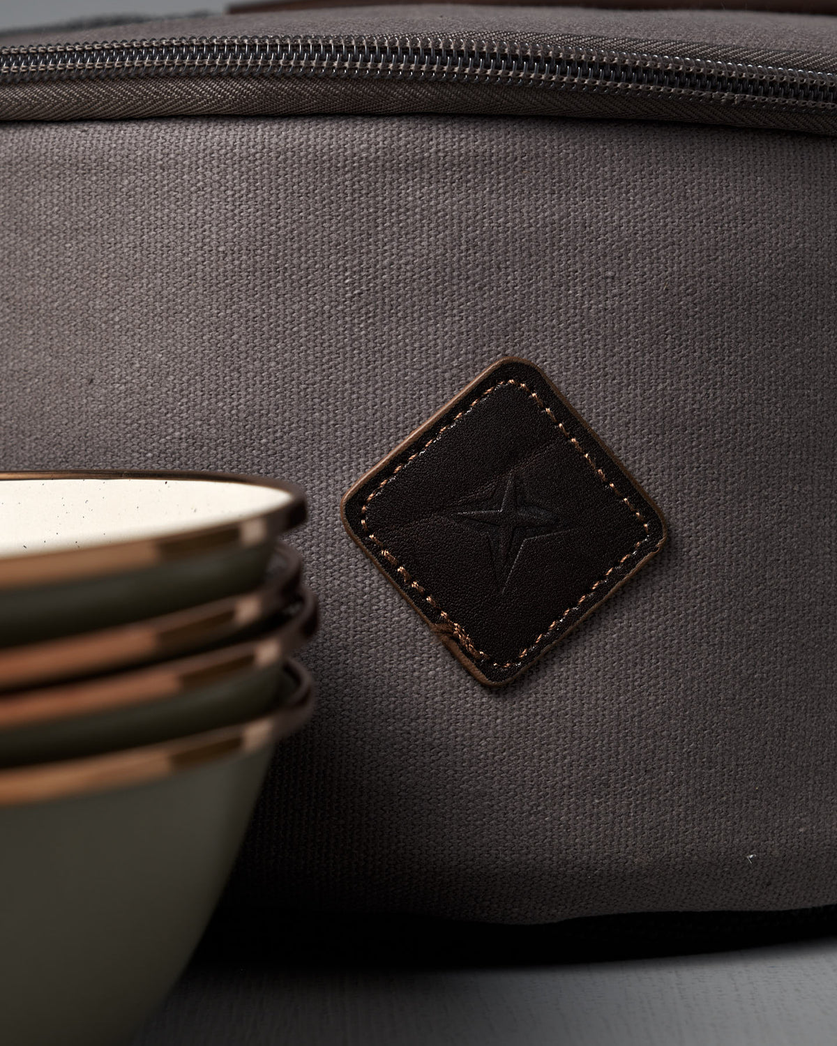 A close-up of stacked ceramic bowls next to the Barebones Padded Utility Storage Bag, crafted from durable cotton canvas and finished with a stitched leather patch featuring a star symbol.