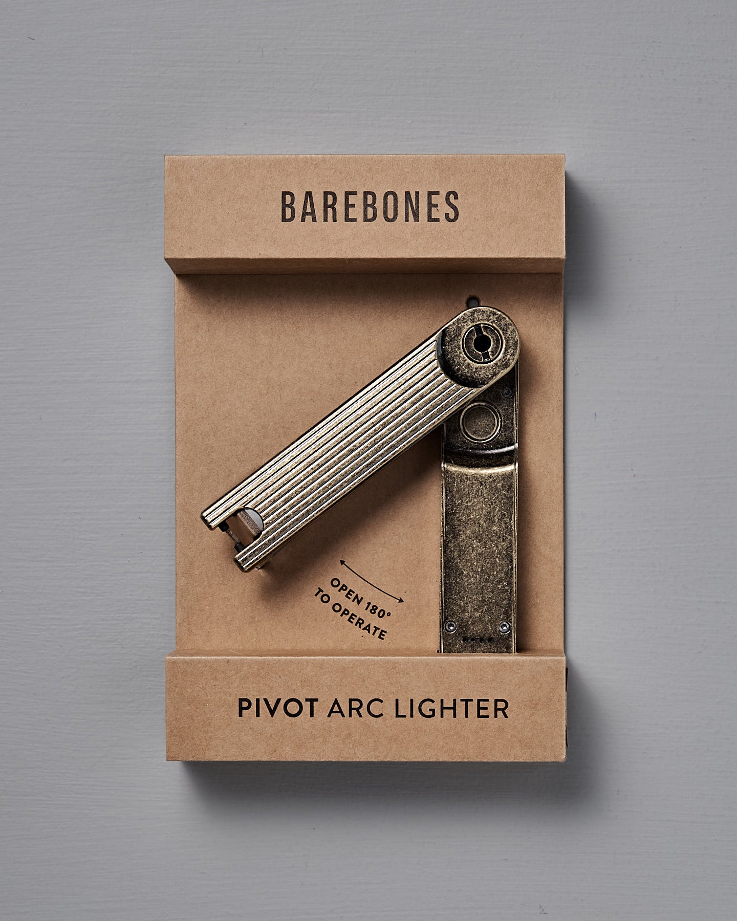 The Barebones Pivot Arc Lighter, featuring a brass finish, is shown in its cardboard packaging against a gray background.