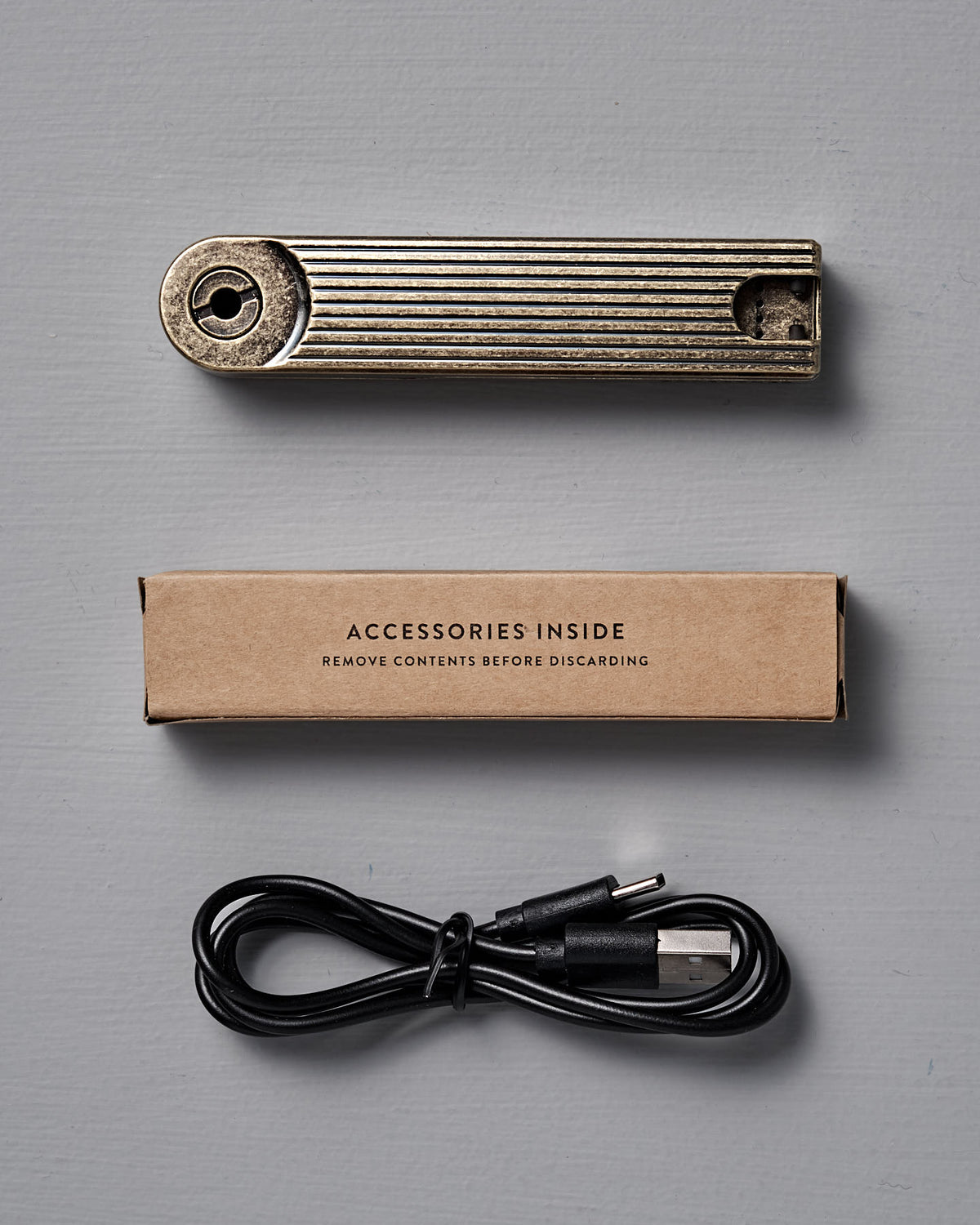 A Barebones Pivot Arc Lighter, cardboard accessories box, and black USB cable are arranged on a gray surface—everything you need for your rechargeable lighter.