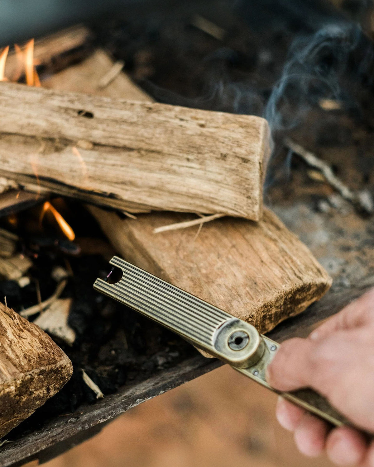 A hand holds the Barebones Pivot Arc Lighter near stacked firewood as a small flame and smoke rise from a fire pit.