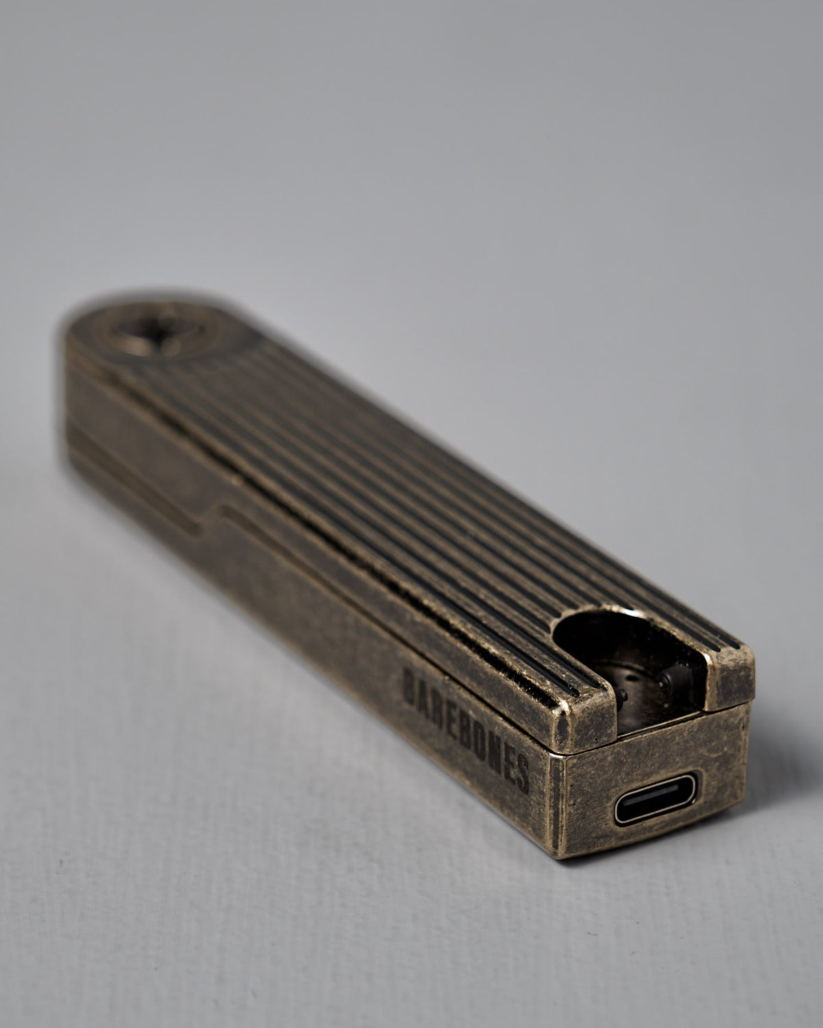 The Barebones Pivot Arc Lighter is a rectangular metal rechargeable plasma lighter with ridged sides, a circular indentation, a USB-C port, and "BAREBONES" engraved on the side.