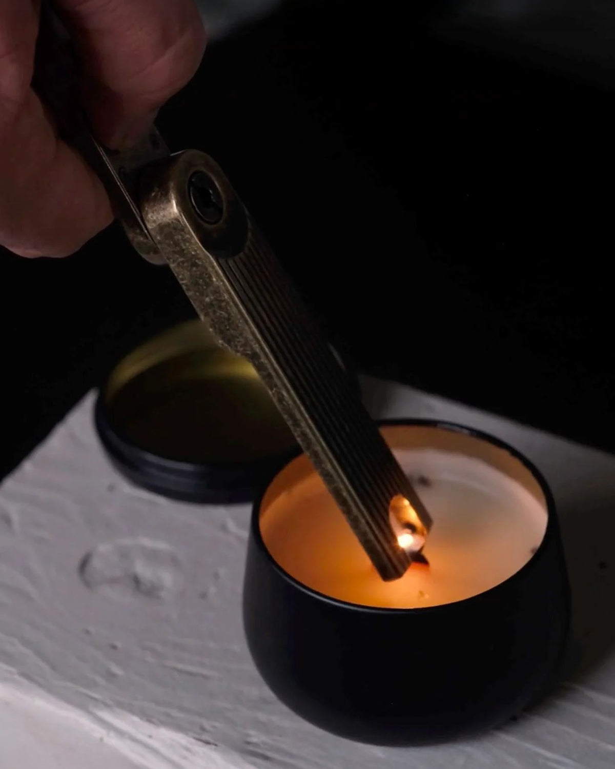 A hand uses the Barebones Pivot Arc Lighter, a windproof electric lighter, to ignite a candle in a black container on a white surface.