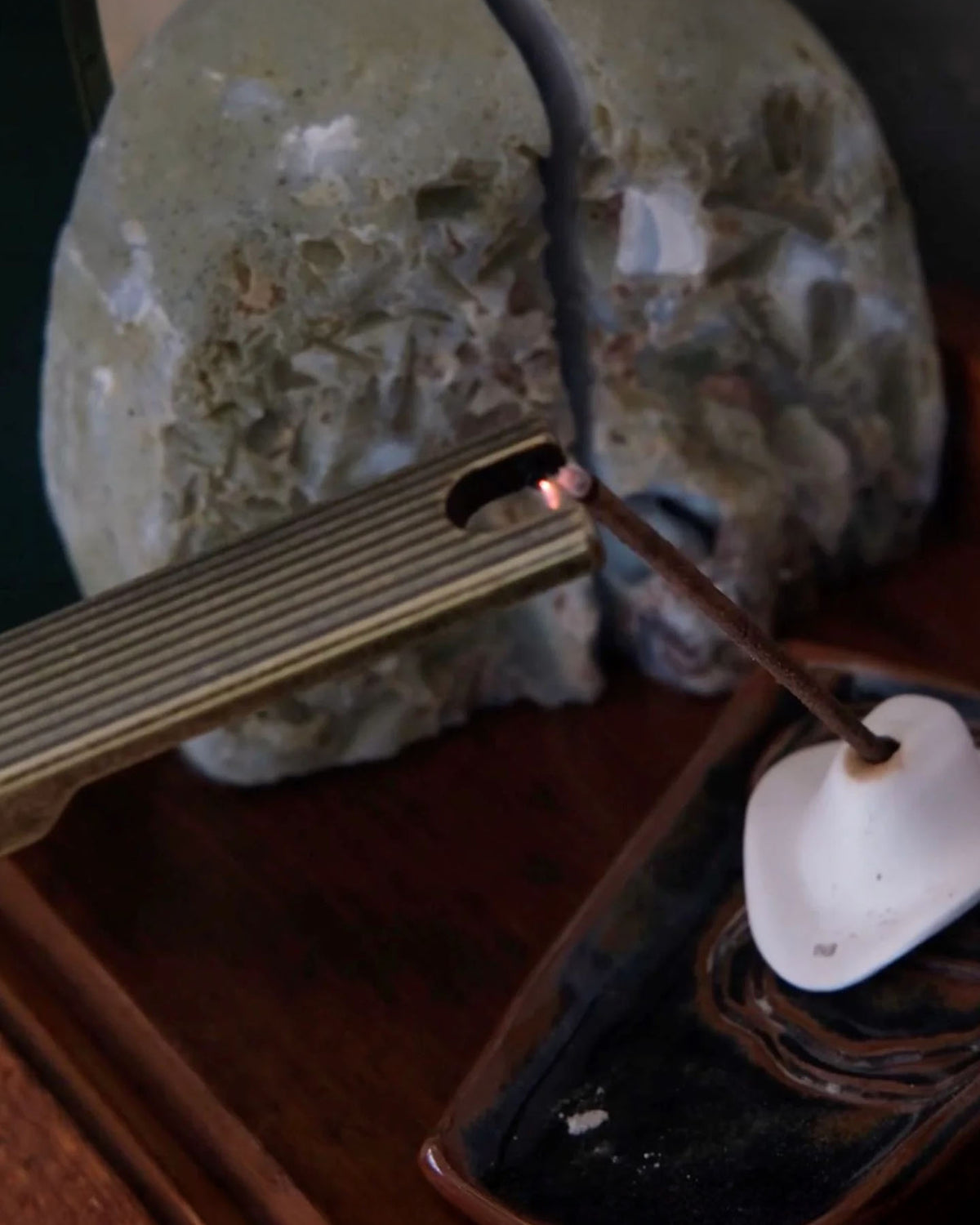 A close-up of a hand using the Barebones Pivot Arc Lighter to ignite an incense stick in a holder on a wooden surface, with a decorative stone object in the background.