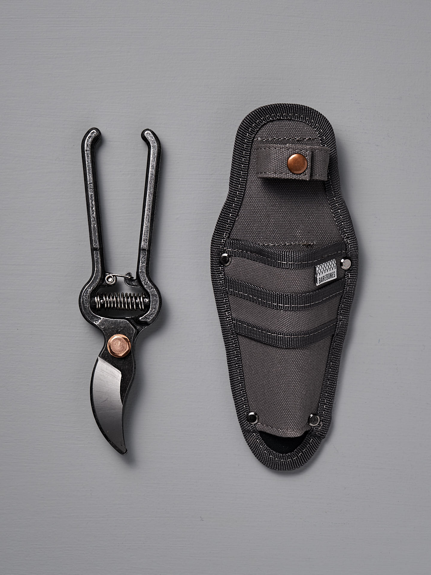 The Barebones Pruner with Sheath, featuring durable steel construction and black handles, is displayed beside its black sheath with button closure and pockets on a gray background.