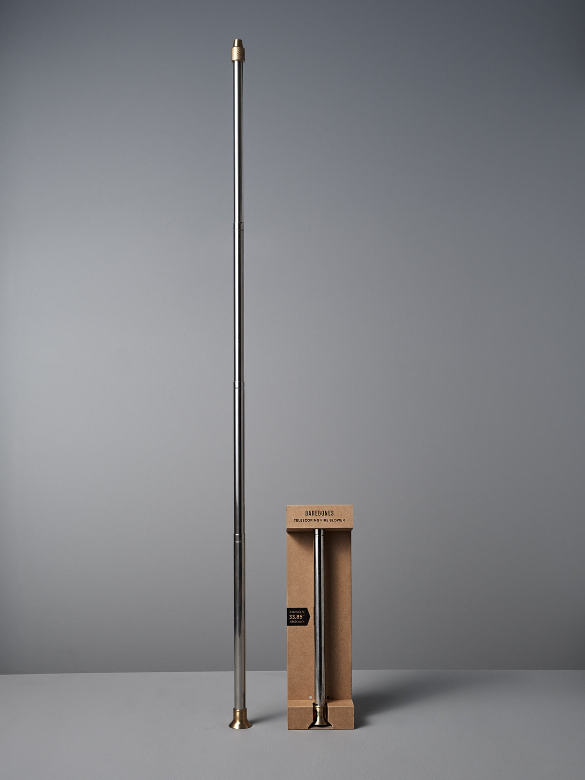 A long, silver Barebones Telescoping Fire Blower with gold-colored tips stands upright beside a shorter, boxed version labeled &quot;Barber’s Pole.