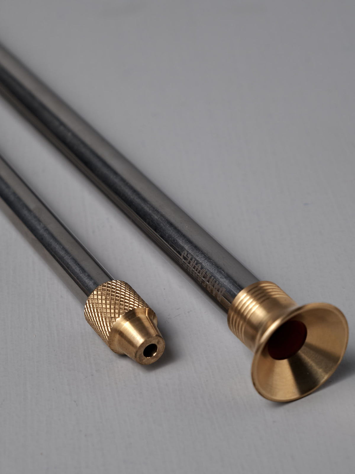 Two metallic rods with brass fittings—one tipped with a knurled cap, the other flared like a trumpet—rest on a light gray surface, highlighting the elegant Barebones Telescoping Fire Blower.