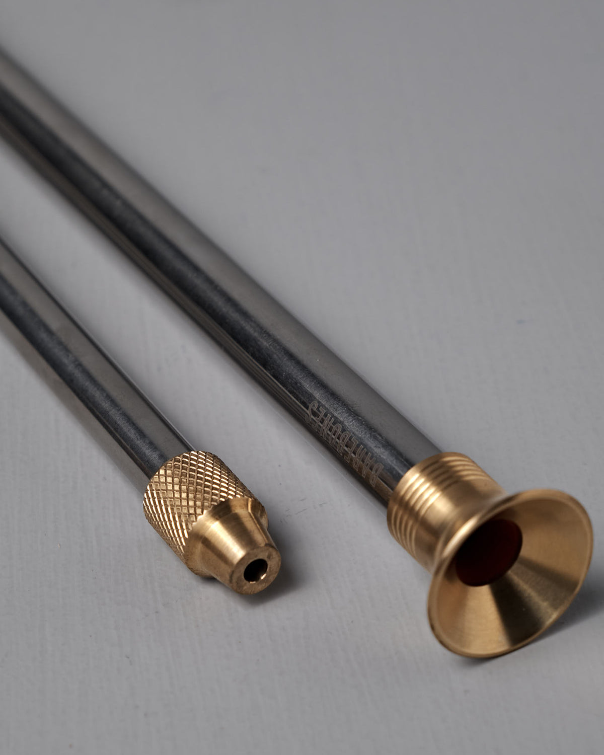 Two metallic rods with brass fittings—one tipped with a knurled cap, the other flared like a trumpet—rest on a light gray surface, highlighting the elegant Barebones Telescoping Fire Blower.