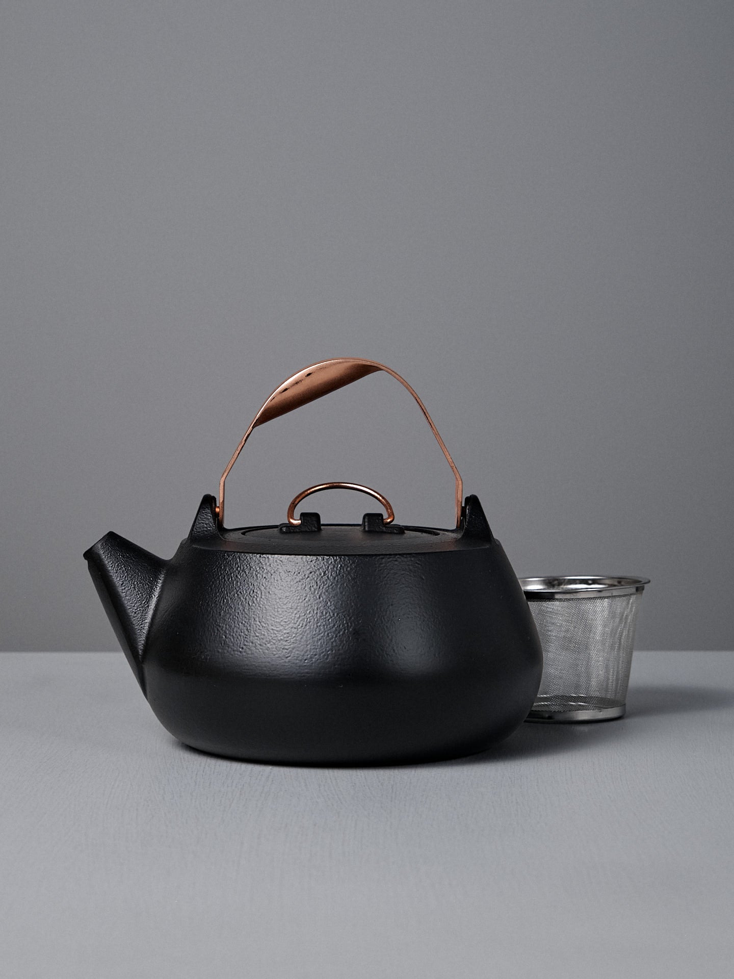 The Cast Iron Teapot (Enamel Lined), featuring a matte black finish and copper handle, offers excellent heat retention and elegance as it sits beside a metal cup on a grey surface.