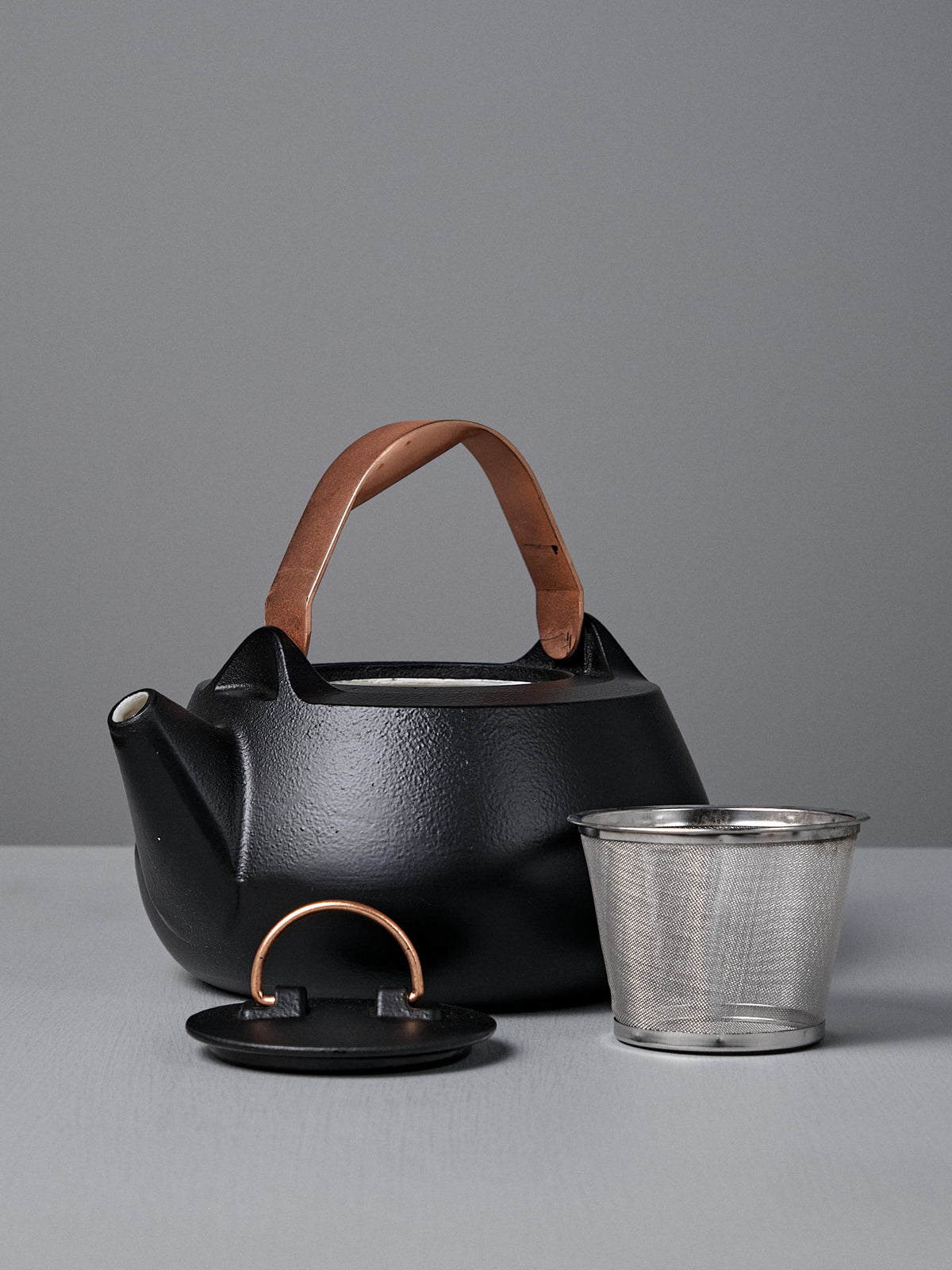 A Cast Iron Teapot (Enamel Lined) with a brown handle is displayed with its lid and stainless steel infuser on a gray surface against a matching gray background.