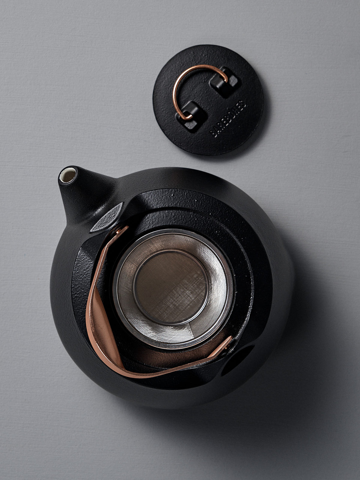 A Cast Iron Teapot (Enamel Lined) in black, shown with its lid off, displays a stainless steel infuser and a copper handle, set on a gray surface.