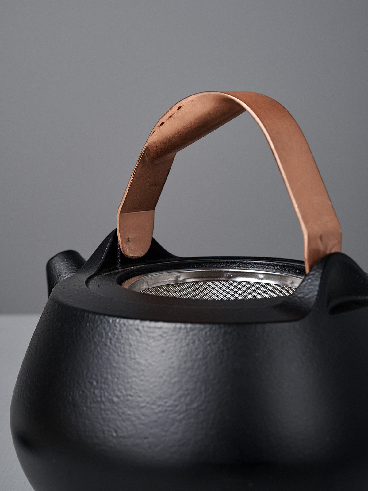 Close-up of the Cast Iron Teapot (Enamel Lined) with superior heat retention, a copper handle, and stainless steel infuser, shown against a plain gray background.