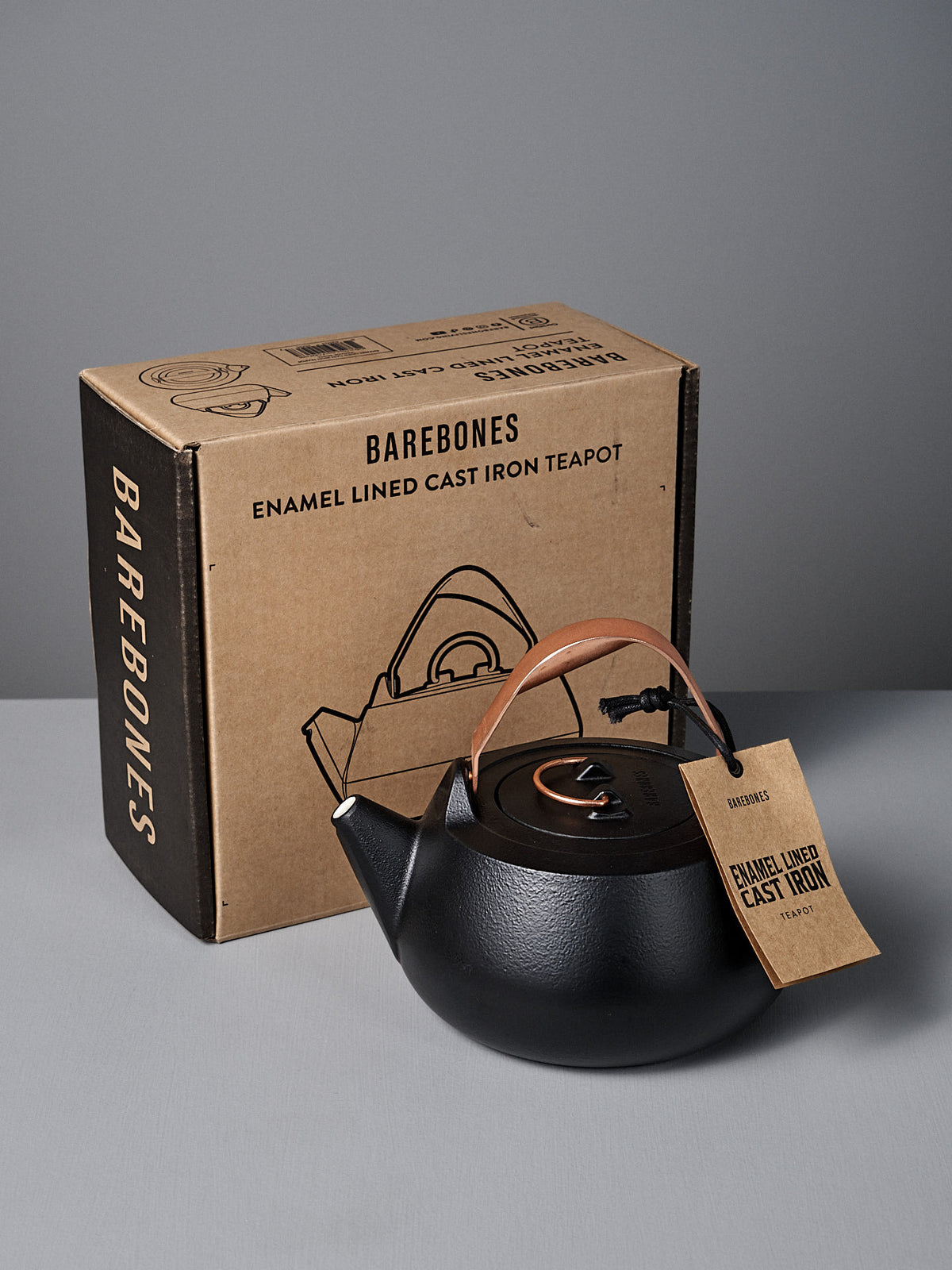 A black Cast Iron Teapot (Enamel Lined) with a brown handle and tag sits before a Barebones box labeled &quot;Enamel Lined Cast Iron Teapot,&quot; known for its excellent heat retention.