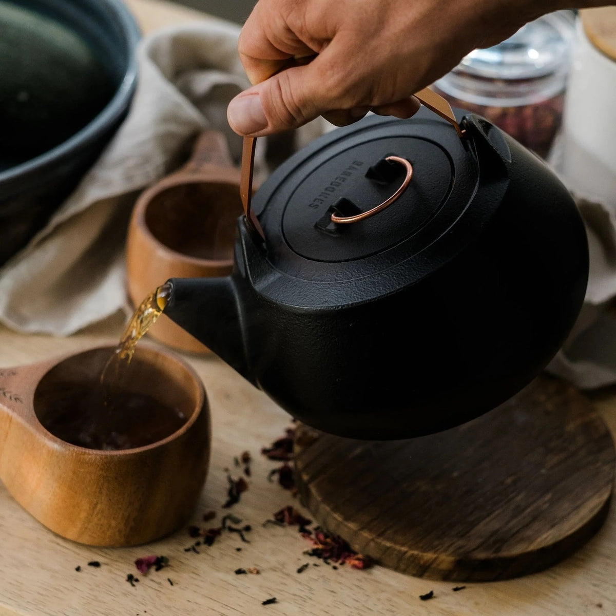 A hand pours tea from a Cast Iron Teapot (Enamel Lined) into a wooden cup on a wooden surface, showcasing the teapot&#39;s superior heat retention. Another cup and loose tea leaves are also visible nearby.