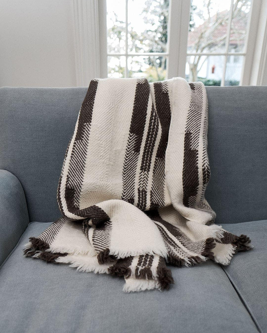 A Rodopska Takan Bulgarian Shevitza Wool Blanket – Cream, made from locally sourced wool, with fringed edges, is draped over a light blue couch in a bright room with large windows in the background.