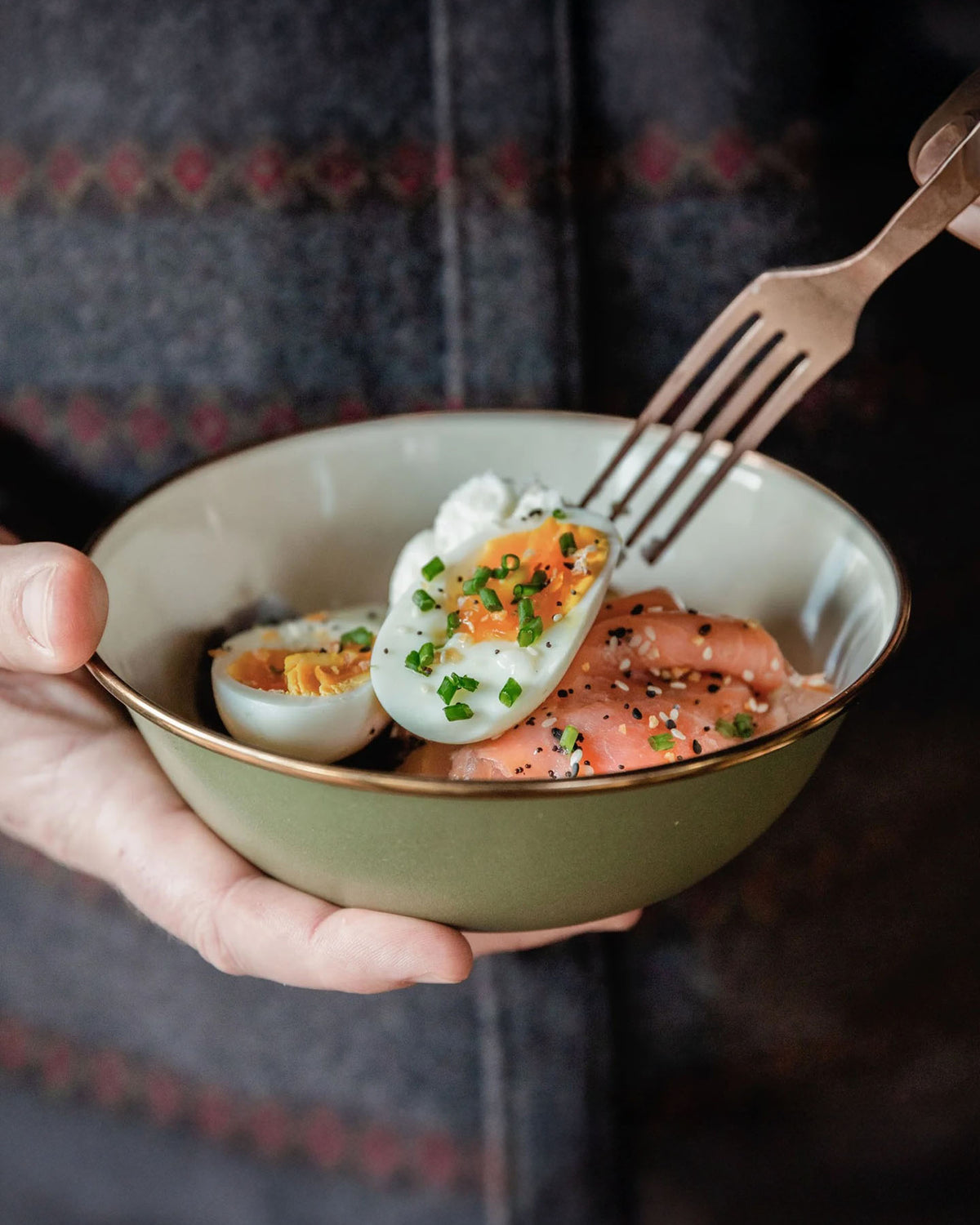 A person uses a fork to hold an Olive Drab Enamel Two-Tone Bowl by Barebones, filled with smoked salmon, half a boiled egg, chives, and cream cheese.