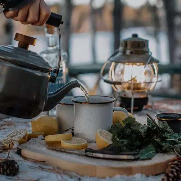 A person pours hot water from a kettle into a white mug on a table with lemons, mint, a lantern, and other items, outdoors near a body of water.