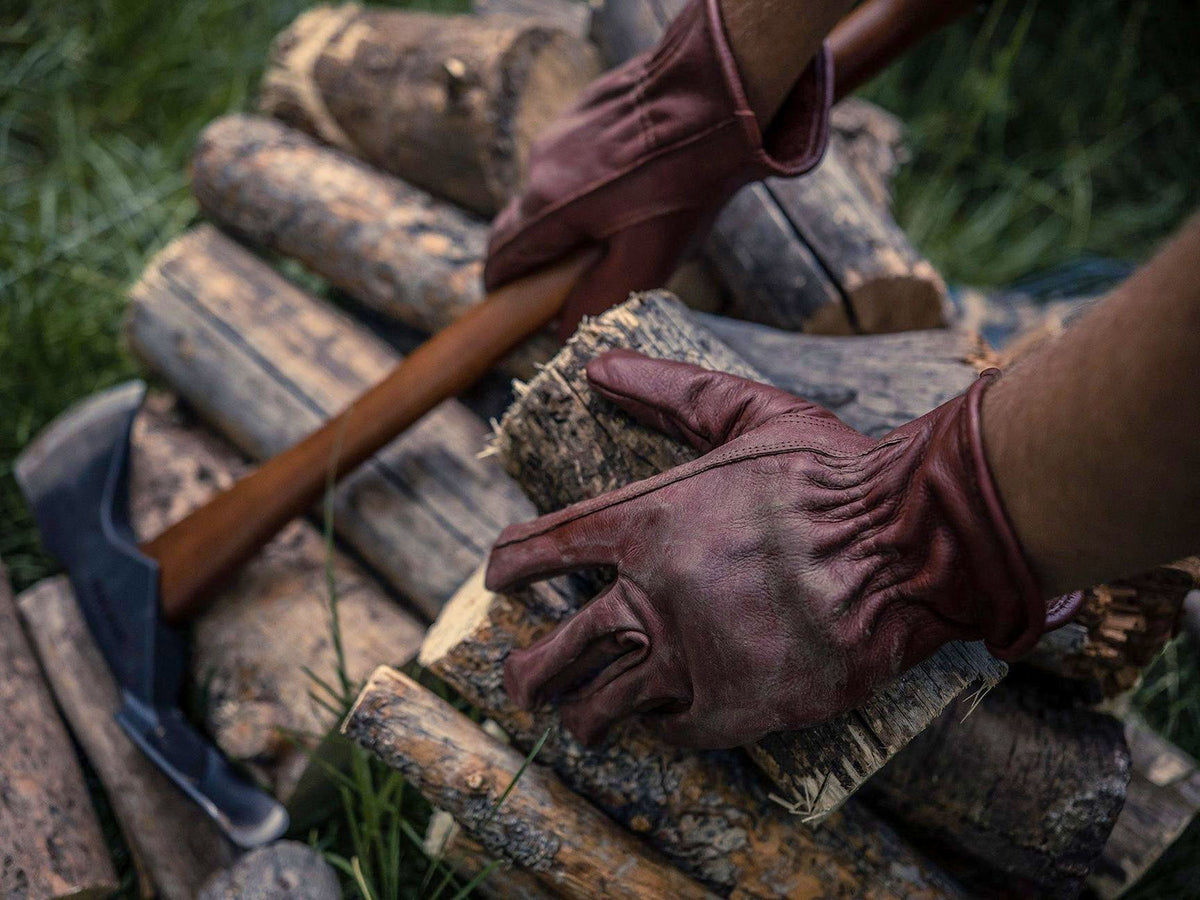 Wearing Barebones&#39; Classic Work Glove – Cognac gloves, a person skillfully manages firewood with an axe on stacked logs, demonstrating outdoor versatility.