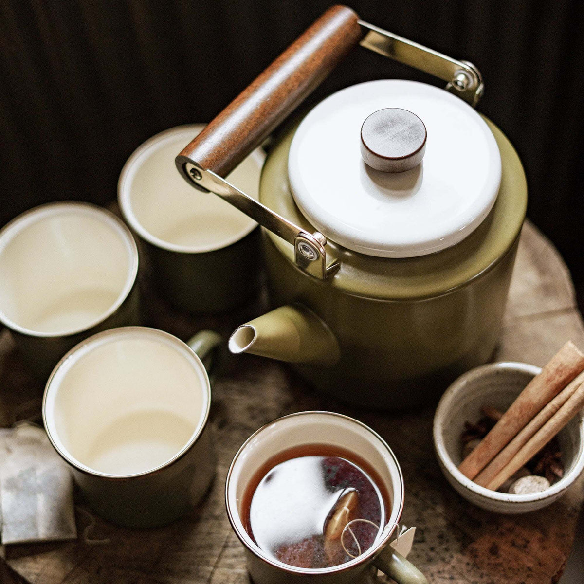 An Enamel Two-Tone Kettle in Olive Drab, crafted by Barebones, with its walnut handle rests next to four complementary cups on a wooden table. One cup is filled with tea, accompanied by a tea bag, a small bowl of spices, and cinnamon sticks nearby.