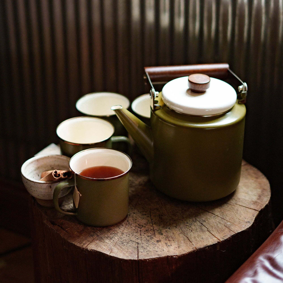 An Olive Drab Enamel Two-Tone Kettle by Barebones, featuring a vintage-inspired design with a walnut handle and white lid, rests on a wooden surface surrounded by three empty mugs and a fourth partially filled with tea.