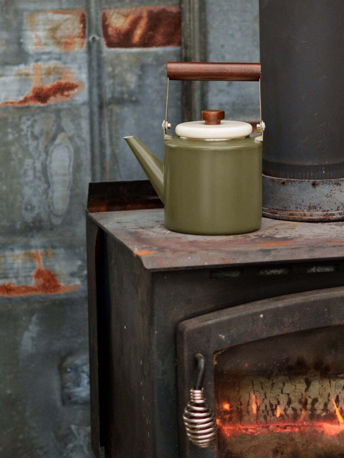 An Enamel Two-Tone Kettle in Olive Drab from Barebones, featuring a vintage-inspired design and walnut handle, sits on a cast iron stove with a rusted background and visible flames inside.