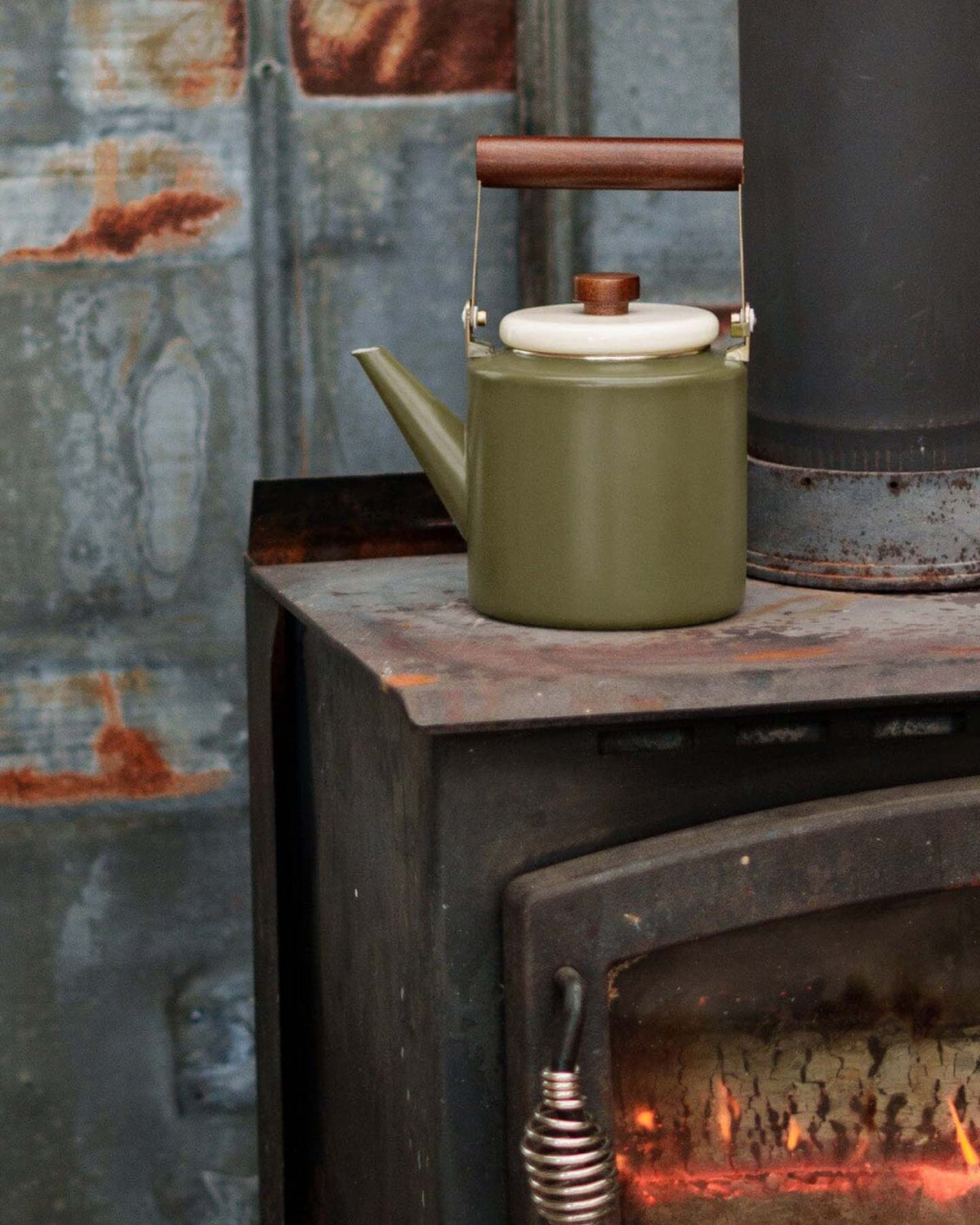 The Enamel Two-Tone Kettle – Olive Drab, featuring a walnut handle, rests atop a black wood-burning stove as the fire inside casts a warm glow.