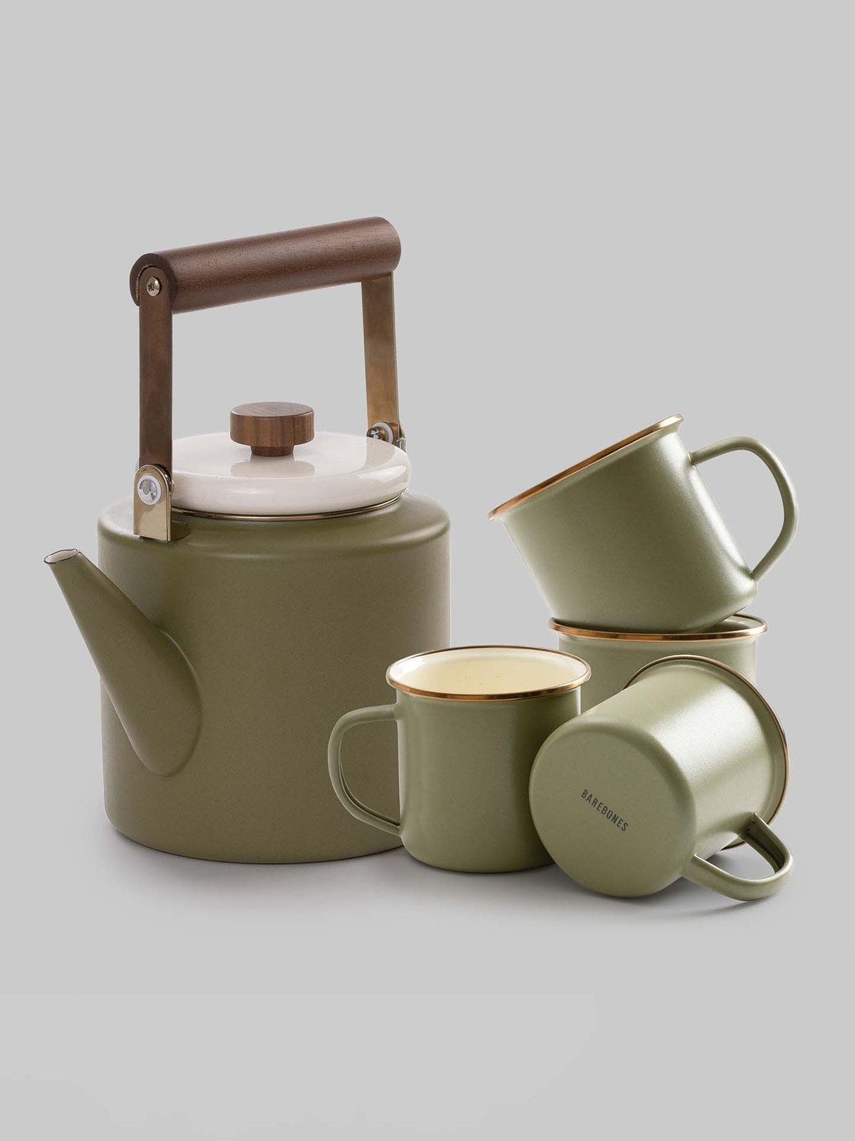 The Enamel Two-Tone Kettle in Olive Drab by Barebones, featuring a retro design with a walnut handle and white lid, is elegantly positioned beside three green mugs adorned with gold rims on a gray background.