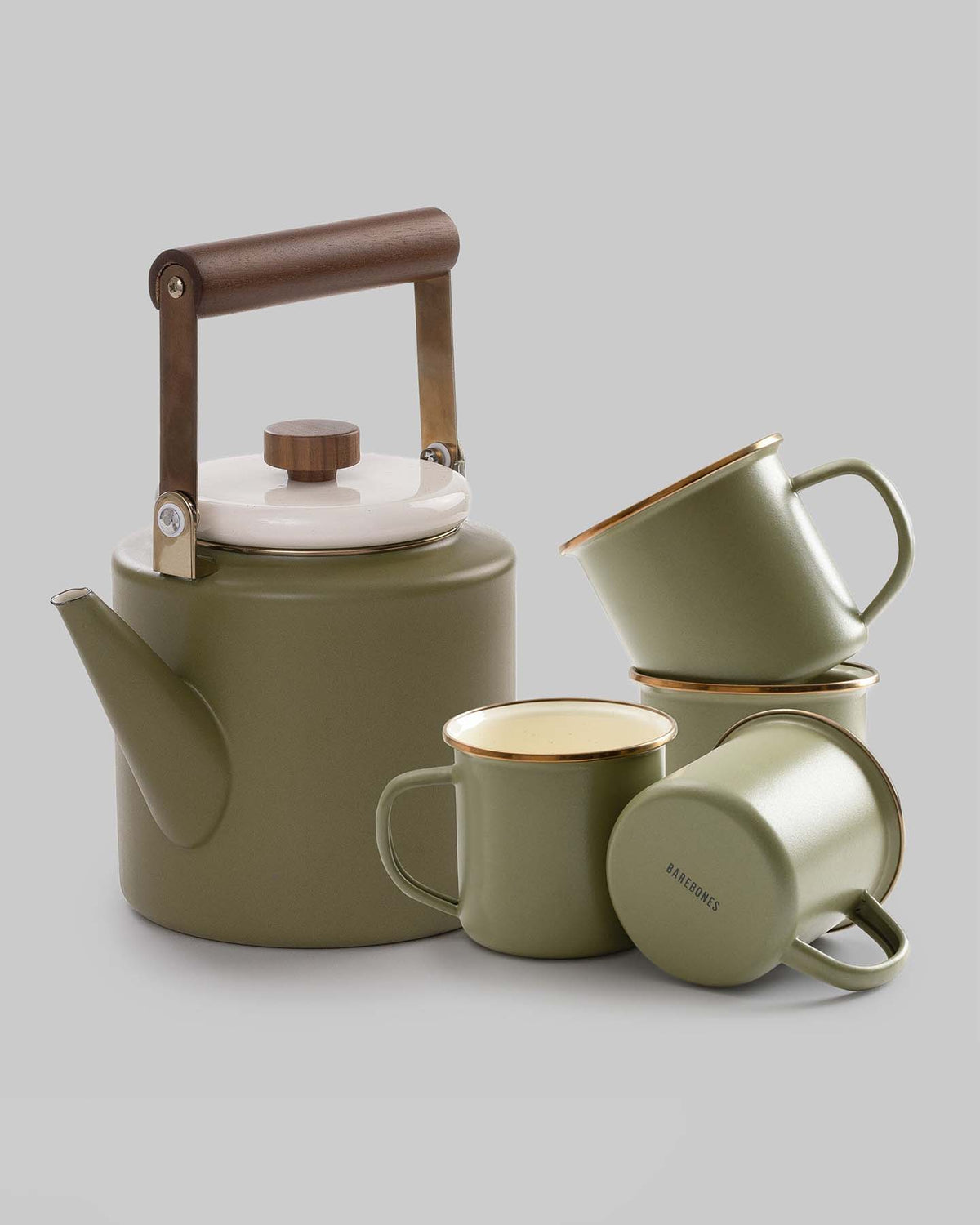 The Enamel Two-Tone Kettle in Olive Drab by Barebones, featuring a retro design with a walnut handle and white lid, is elegantly positioned beside three green mugs adorned with gold rims on a gray background.