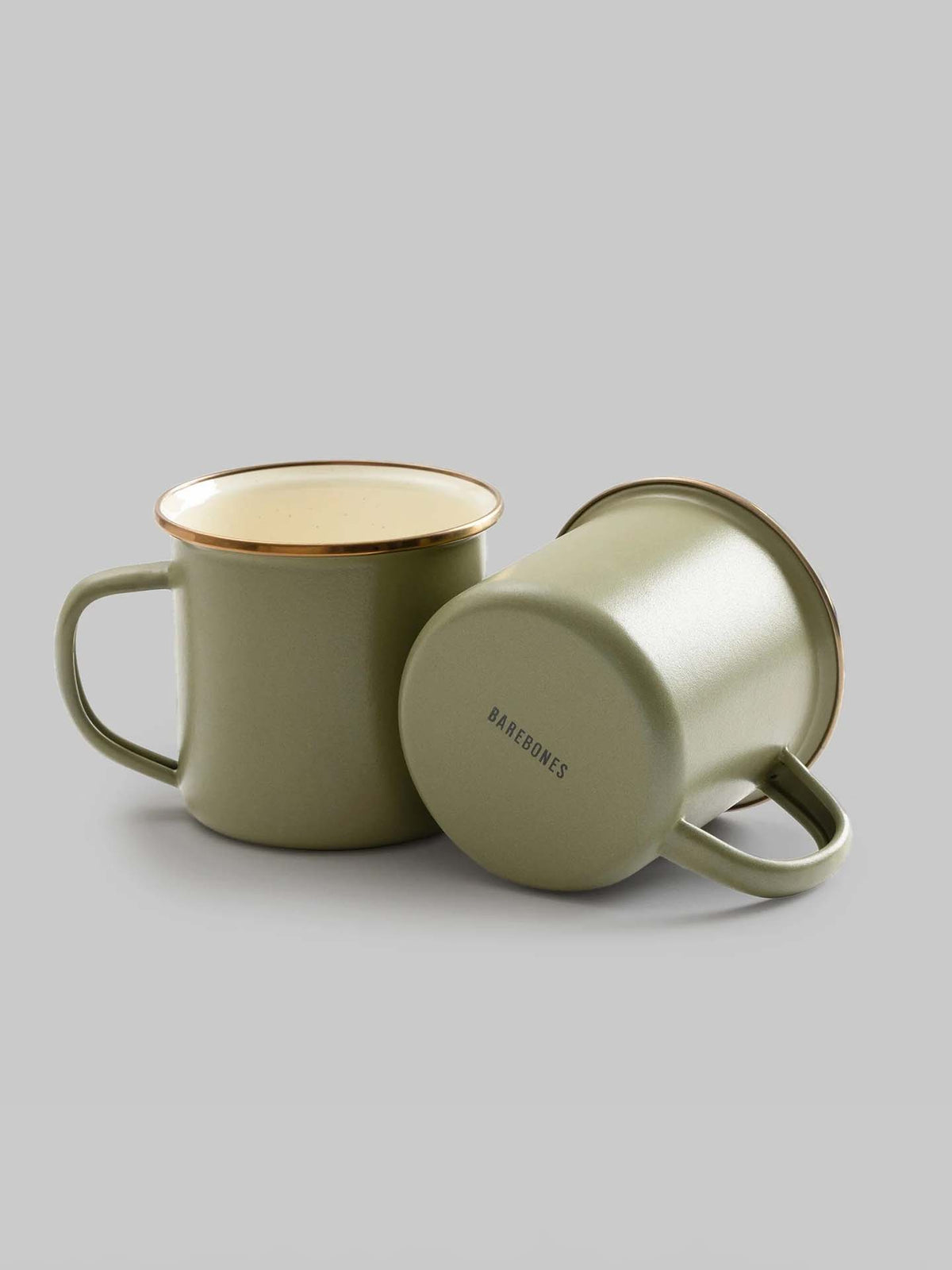 The Enamel Two-Tone Mugs in Olive Drab by Barebones, showcasing a vintage-inspired design, are set against a gray background—one standing upright and the other playfully resting on its side.