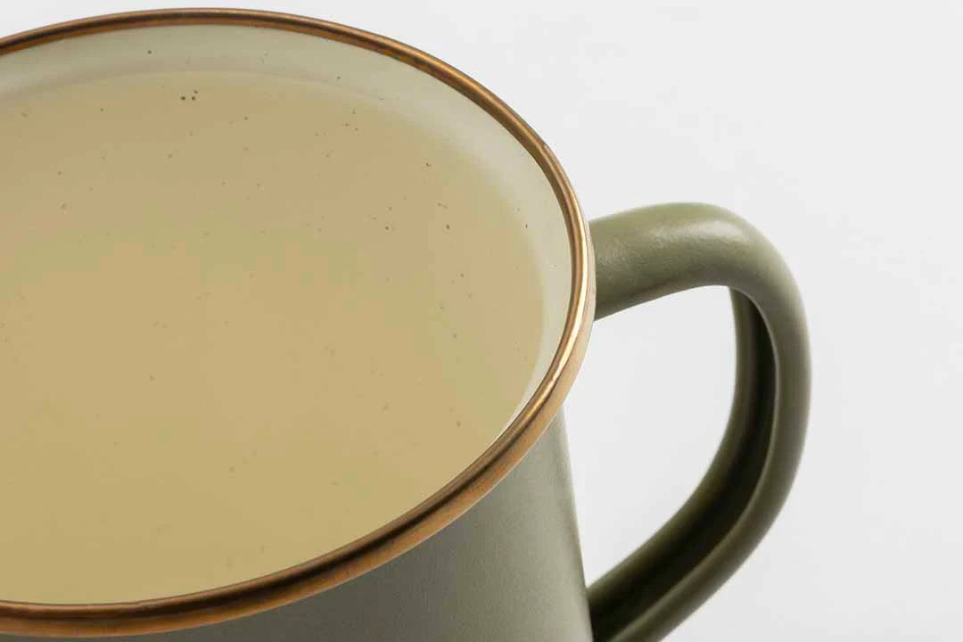 Close-up of an olive drab Barebones Enamel Two-Tone Mug with a beige liquid, viewed at a slight angle against a white background, highlighting the charm of classic enamelware.