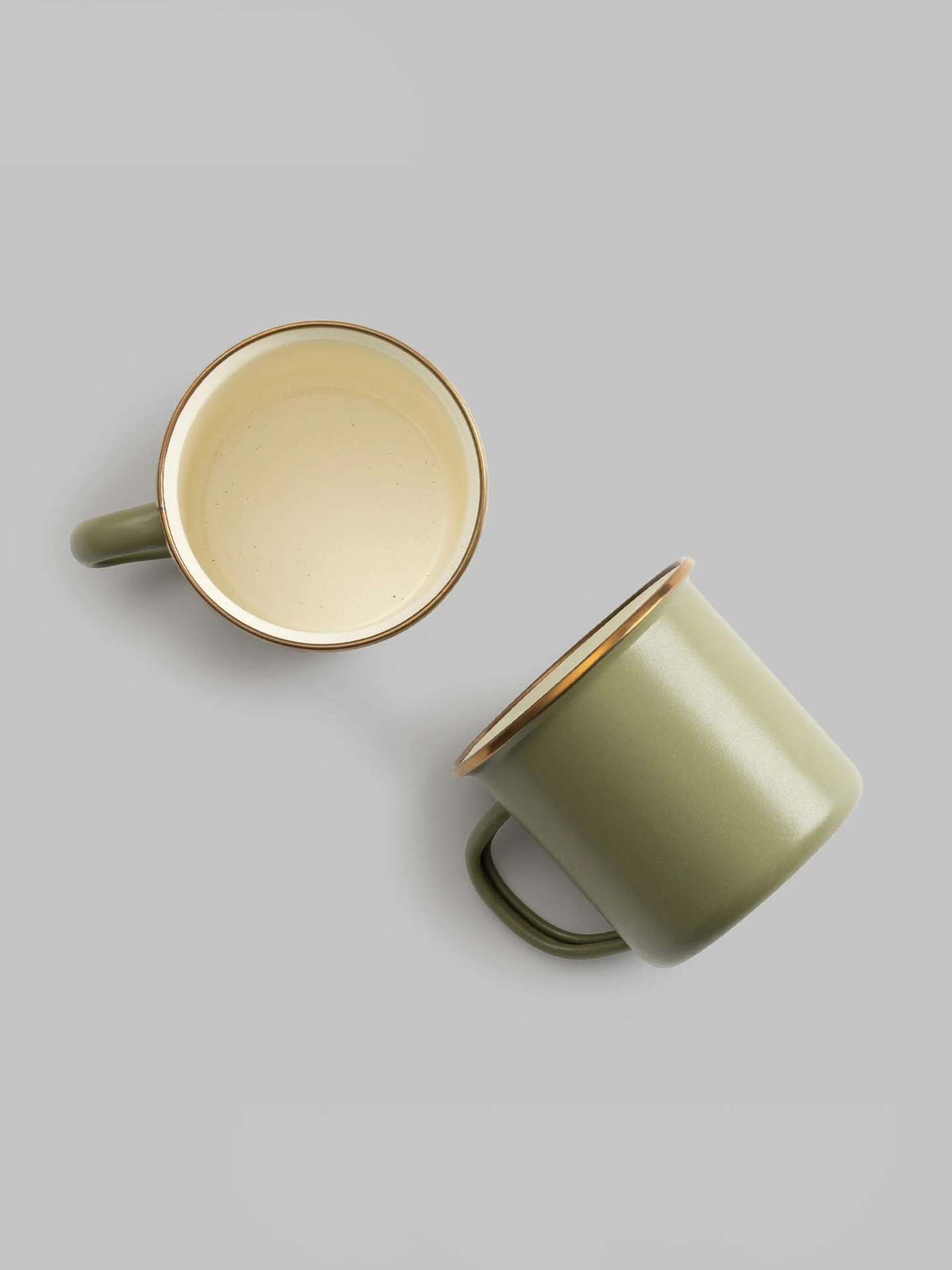 The Enamel Two-Tone Mug set in Olive Drab by Barebones, featuring two vintage-inspired mugs with beige interiors, is artfully displayed against a light gray background, with one standing upright and the other lying on its side.