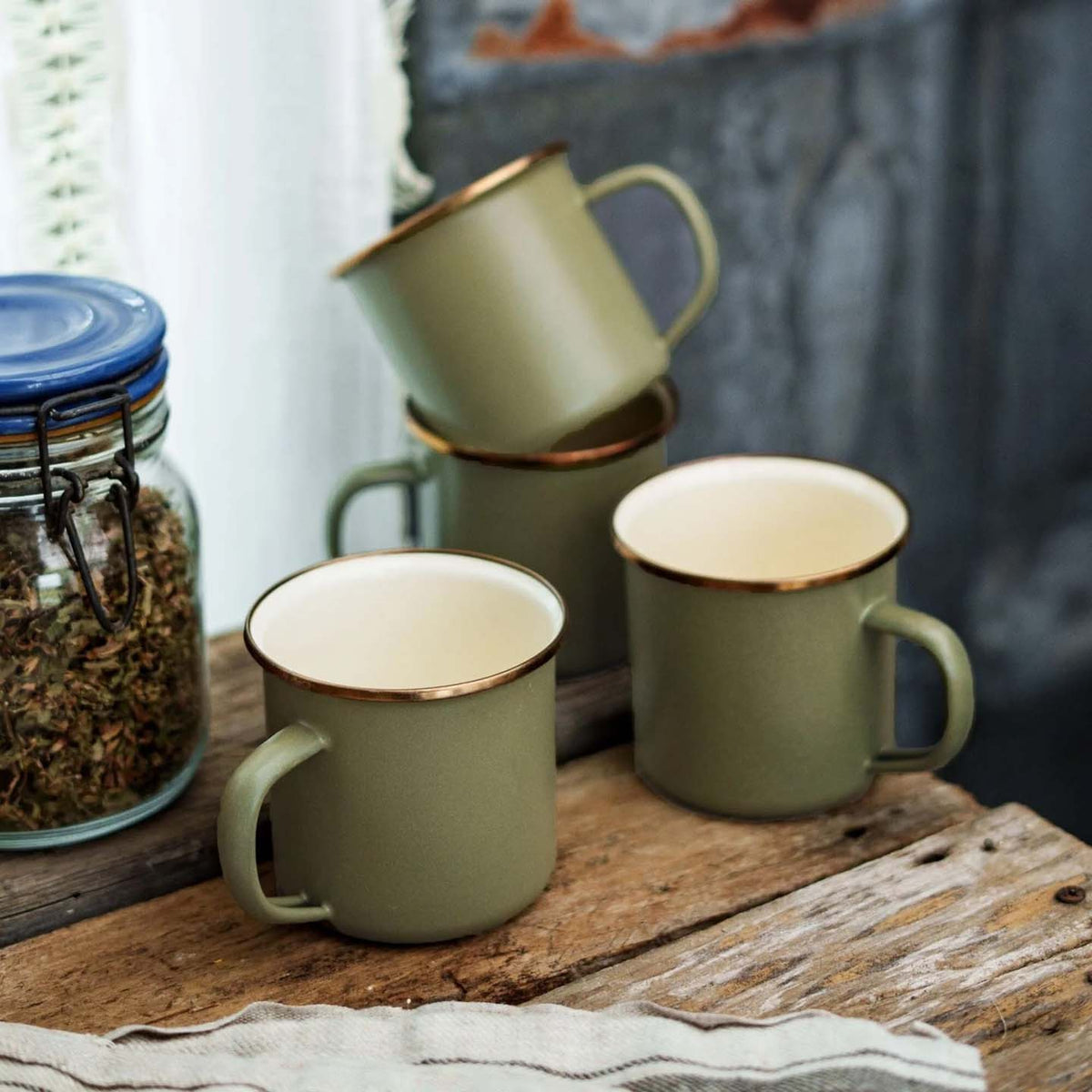 A set of four Enamel Two-Tone Mugs in Olive Drab from the brand Barebones, featuring a vintage-inspired design and gold rims, are neatly stacked on a wooden surface next to a jar filled with herbs.