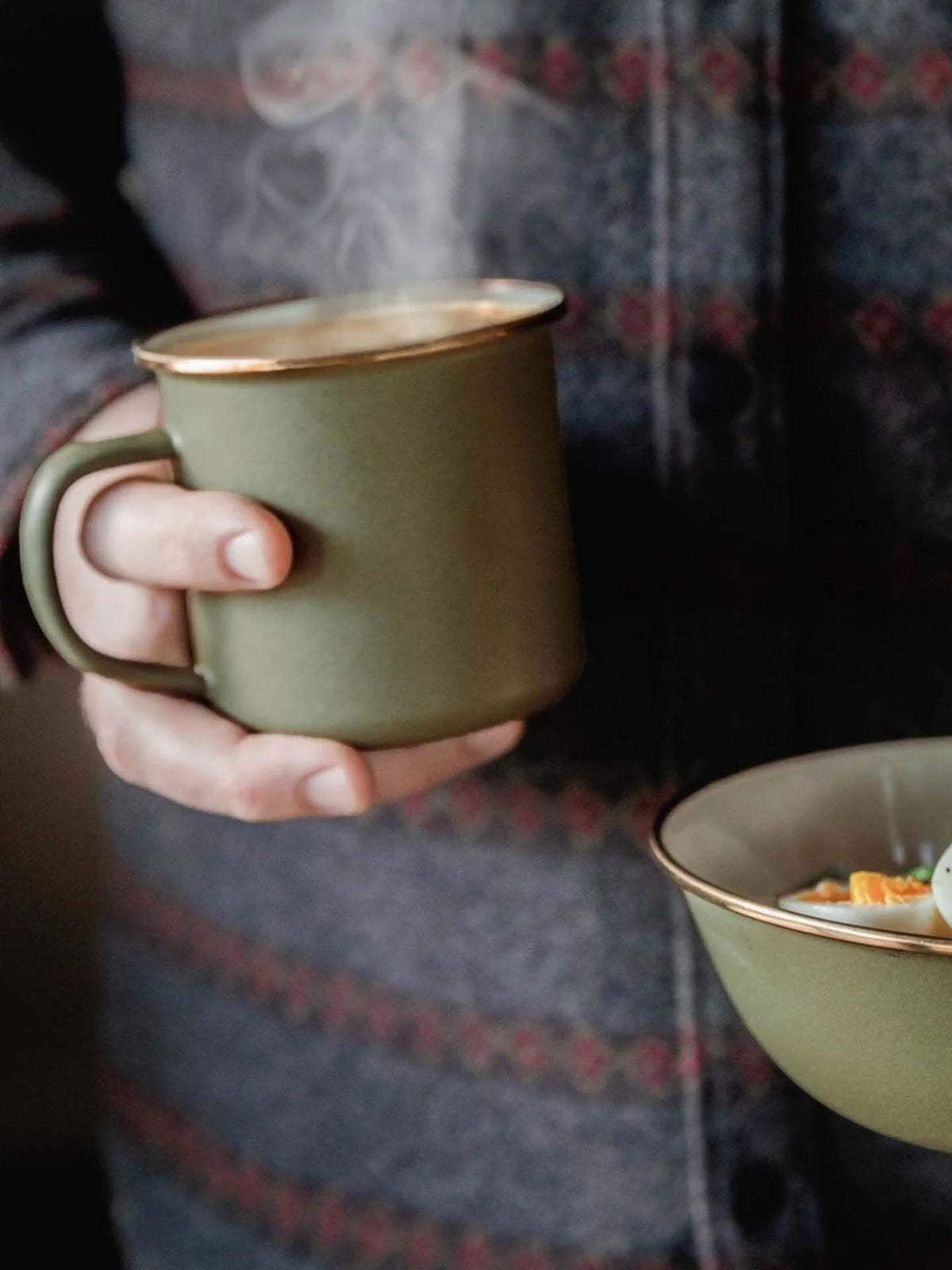 A person holds a steaming Barebones Enamel Two-Tone Mug in Olive Drab and a matching bowl, both featuring a vintage-inspired design, while wearing a patterned jacket.