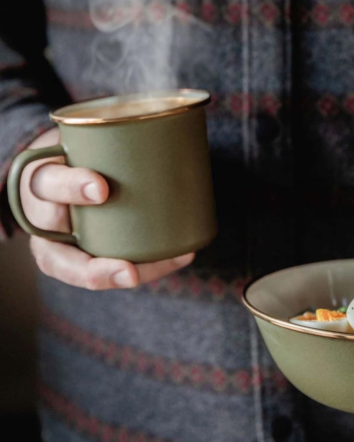 A person holds a steaming Barebones Enamel Two-Tone Mug in Olive Drab and a matching bowl, both featuring a vintage-inspired design, while wearing a patterned jacket.