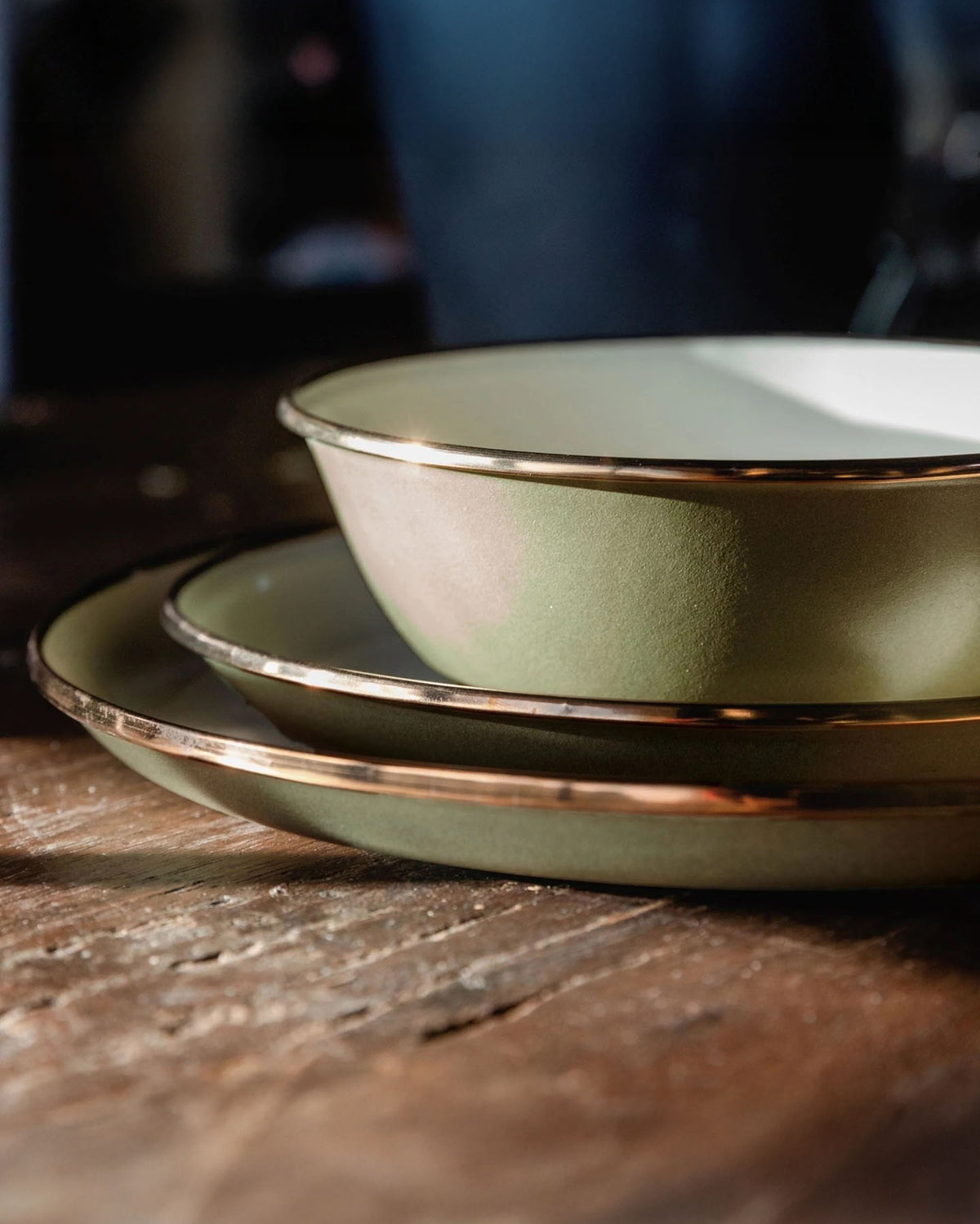 A set of Barebones Enamel Two-Tone Deep Plates in Olive Drab rests on a wooden table, illuminated softly. Their vintage-inspired design exudes timeless elegance, inviting you to dine with charm and grace.
