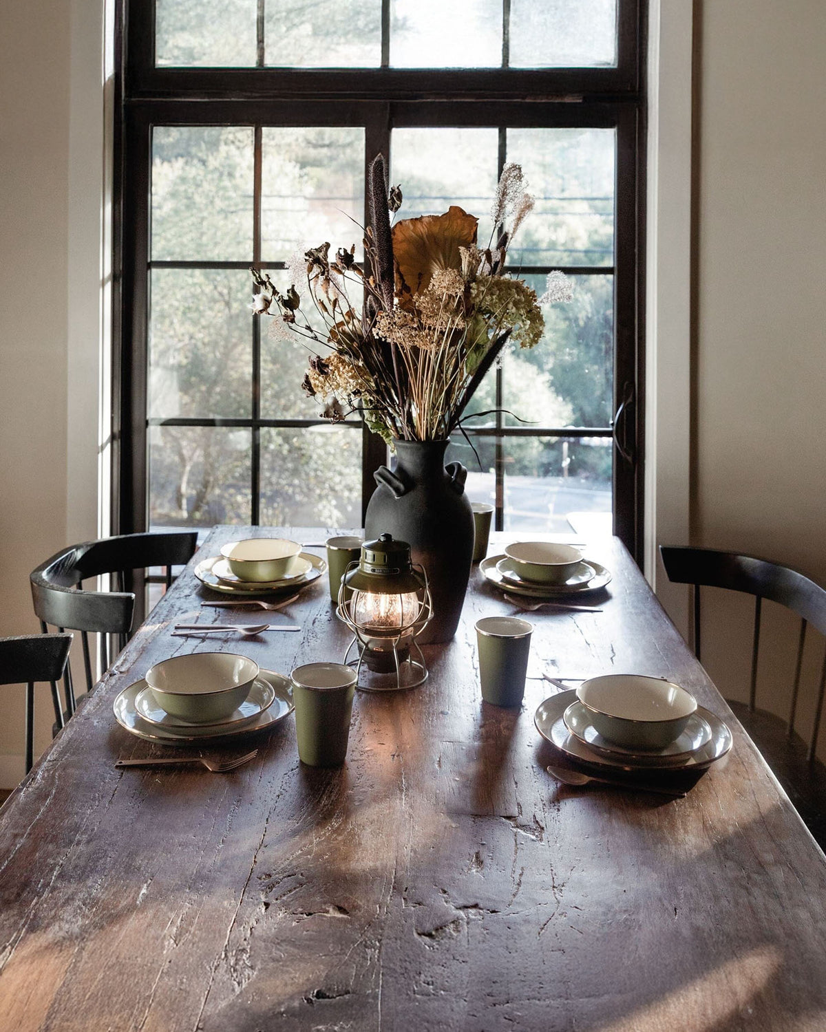 A Barebones dining set featuring the Enamel Two-Tone Deep Plate – Olive Drab, bowls, and cups surrounds a lantern. A vase with dried flowers sits in the center as sunlight from a large window highlights the vintage-inspired decor.