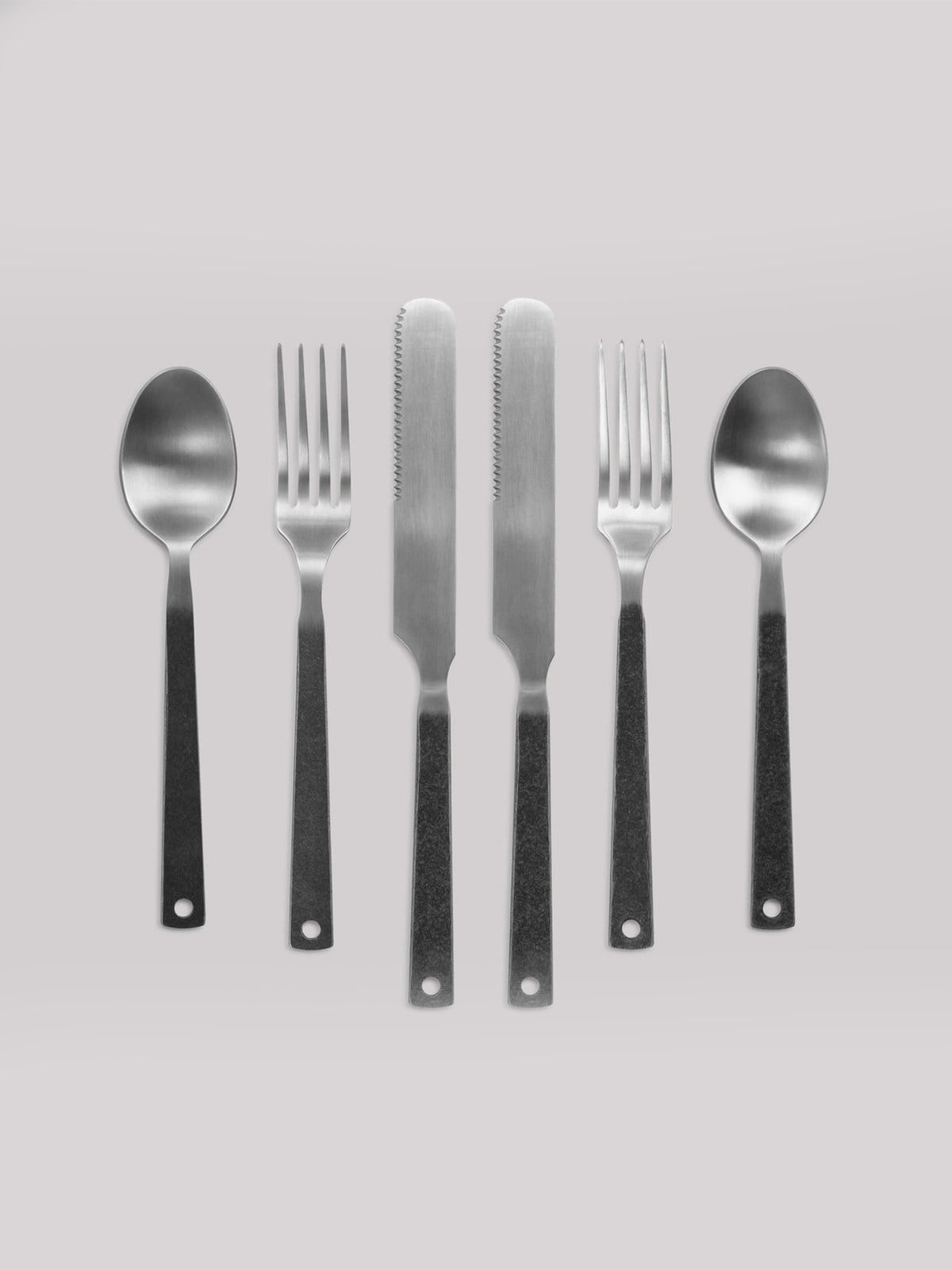 The Barebones Flatware Cutlery set displays two spoons, two forks, and two serrated knives with black handles in alternating order on a light gray background, highlighting its minimalist design.