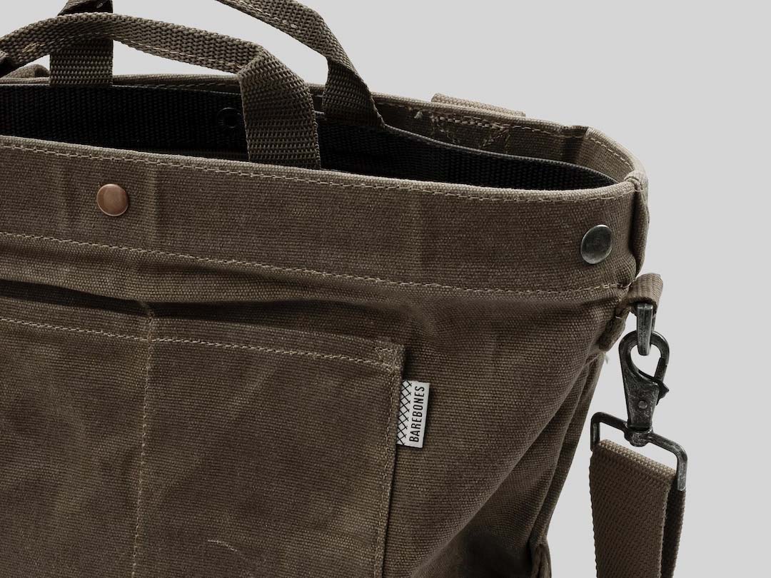 Here&#39;s a close-up of the Gathering Bag – Khaki from Barebones, made from paraffin waxed canvas. It features a strap and metal clip, with a vintage-inspired design that includes a small fabric label on one side and two button details near the opening.