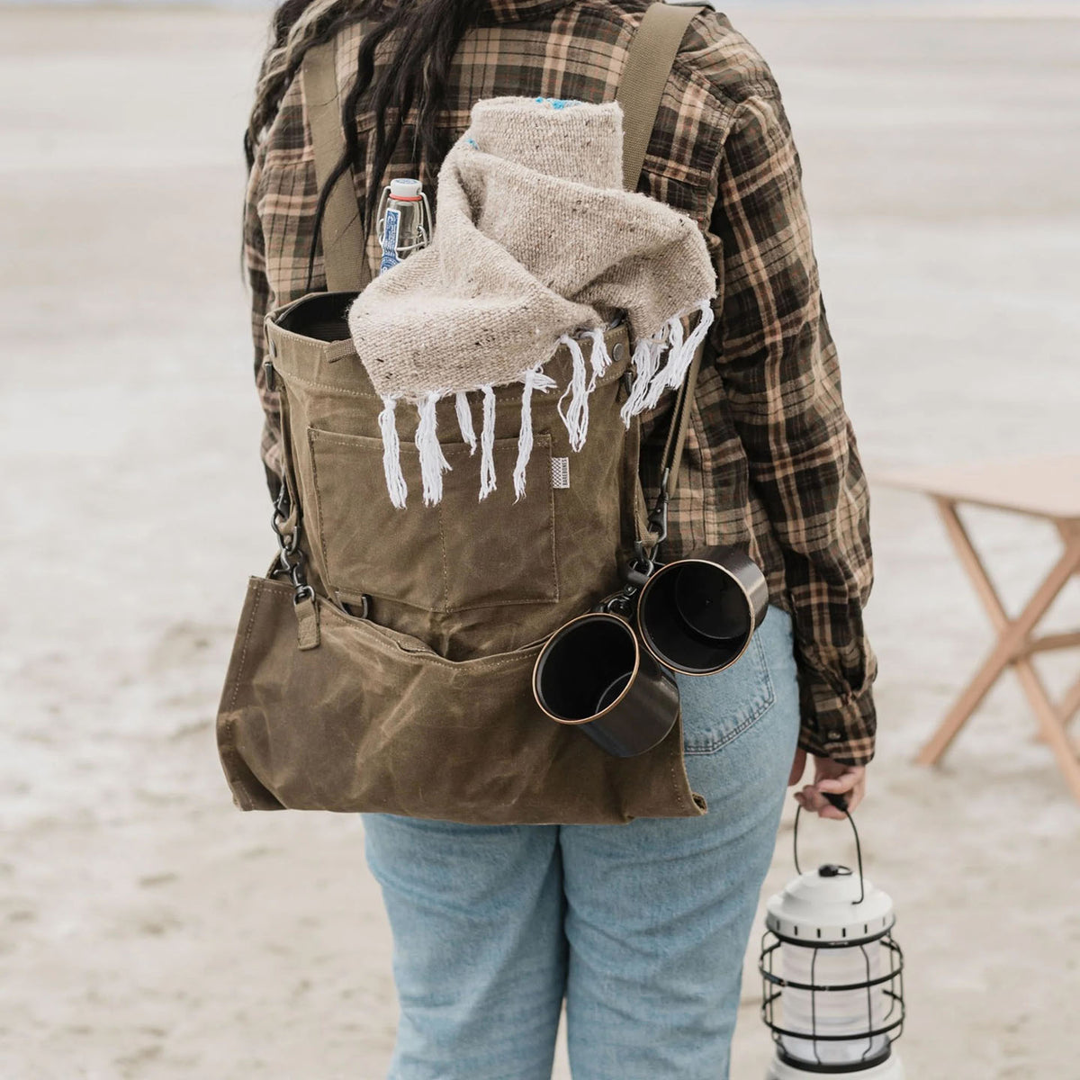 Wearing a plaid shirt and jeans, an individual carries the Barebones Gathering Bag in khaki, filled with a bottle, cloth, and mugs. They hold a lantern in their right hand while standing on a sandy beach beside a wooden chair. The vintage-inspired backpack enhances their rustic style.