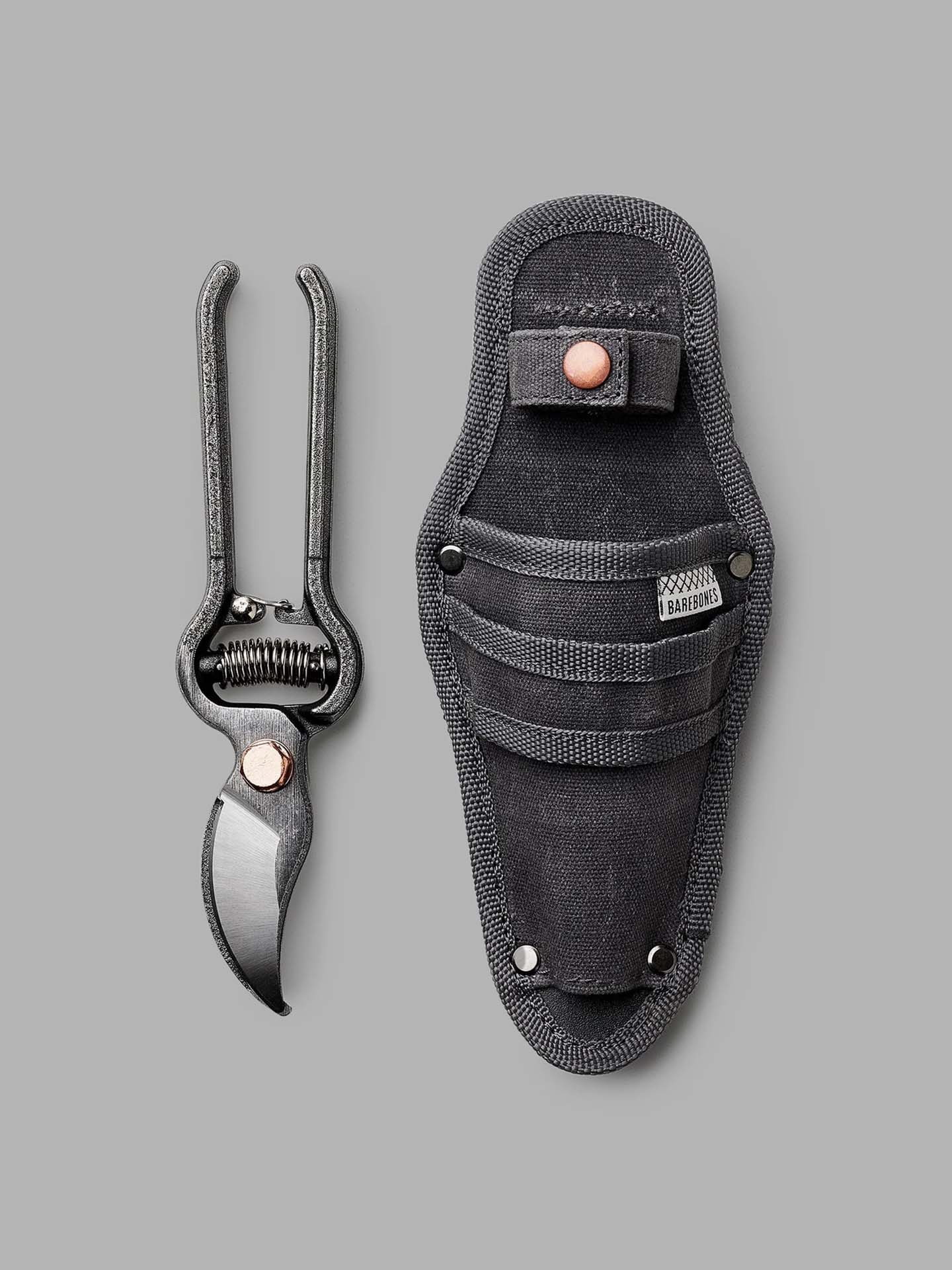 A pair of ultra-durable steel Barebones pruners with a curved blade, in sleek black, is placed alongside their custom sheath on a gray surface.