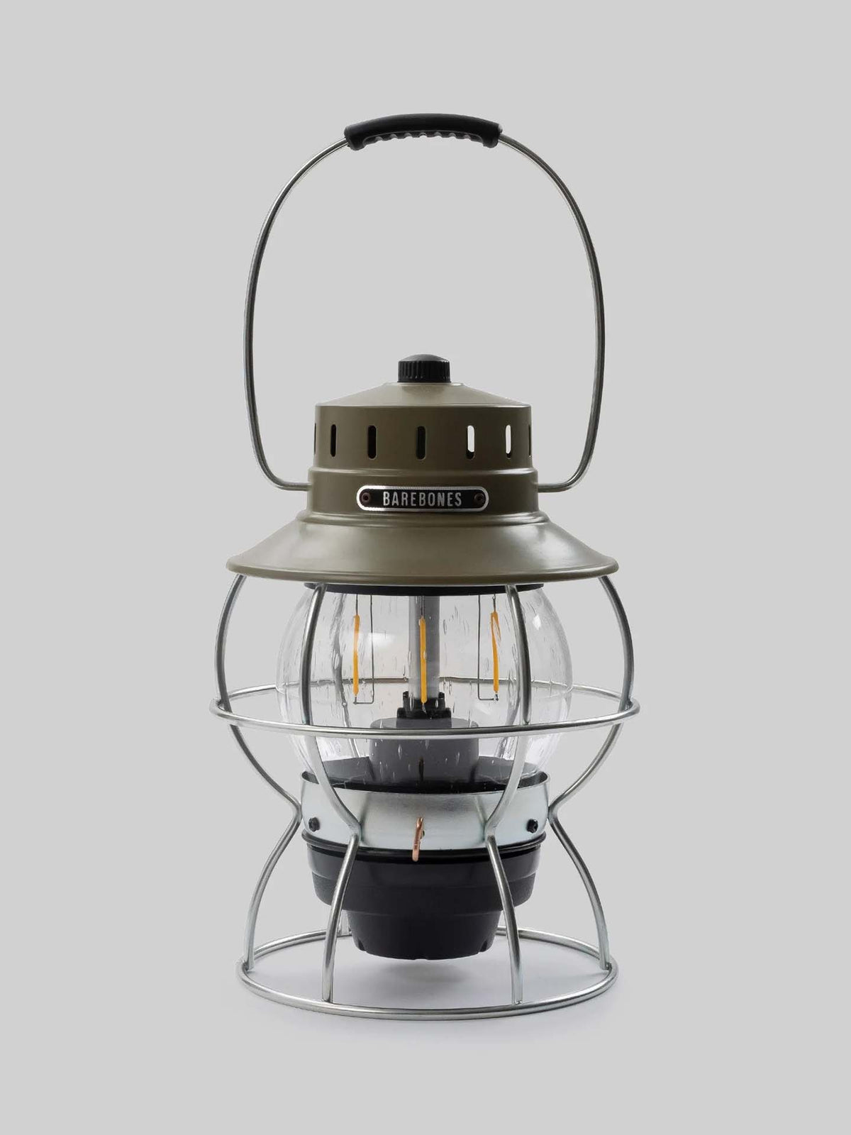 The Barebones Railroad Lantern in Olive Drab features a green and silver design with a clear dome, wire protection, and a top handle. It includes an Edison-style LED bulb that offers a vintage glow.