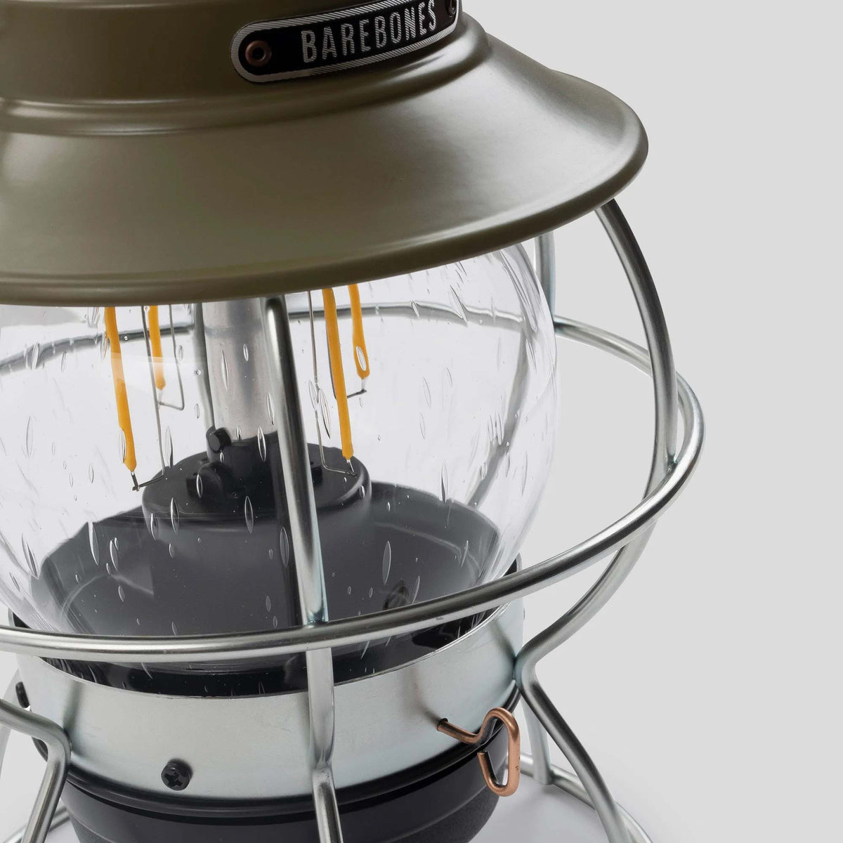 A close-up of the Barebones Railroad Lantern in Olive Drab, showcasing its green and silver exterior with a glass cover and an Edison-style LED bulb. Its design evokes the classic Union Pacific lantern, enhanced with a rechargeable battery for contemporary convenience, all set against a plain background.