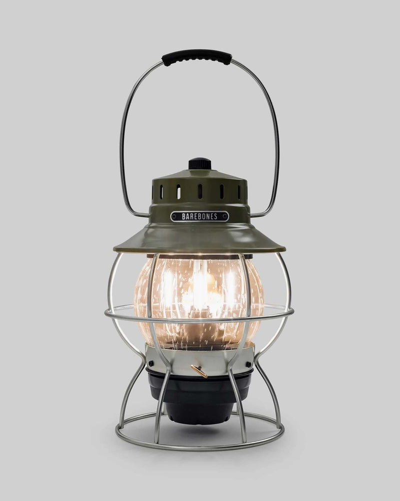 The Railroad Lantern – Olive Drab by Barebones, showcasing an Edison-style LED bulb and a transparent glass globe with a vintage-style olive green top, is set against a plain gray background.