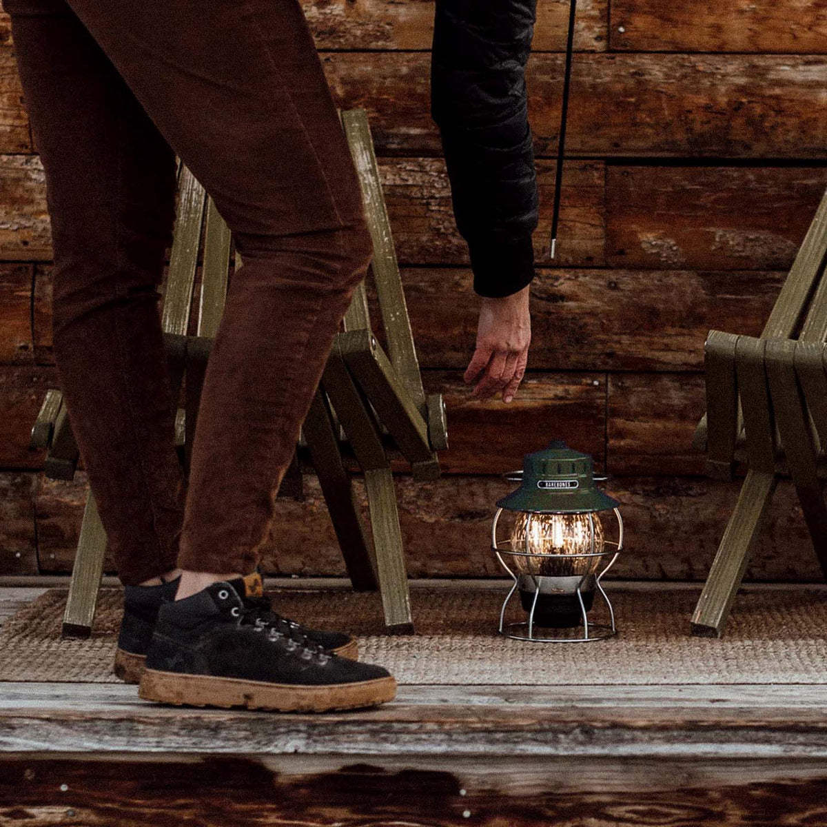 An individual gently sets a Railroad Lantern – Olive Drab by Barebones atop a rug between two wooden chairs, its glow accentuated by an Edison-style LED bulb operated by a rechargeable battery.