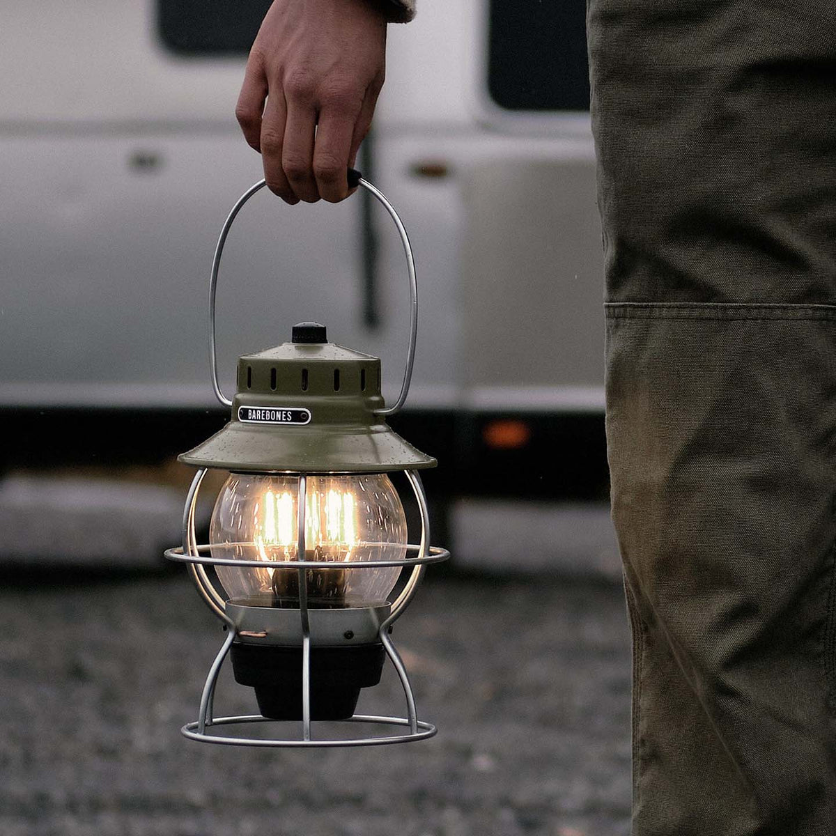 A person holds a lit Barebones Railroad Lantern in Olive Drab, featuring an Edison-style LED bulb, outdoors against a beautifully blurred background.