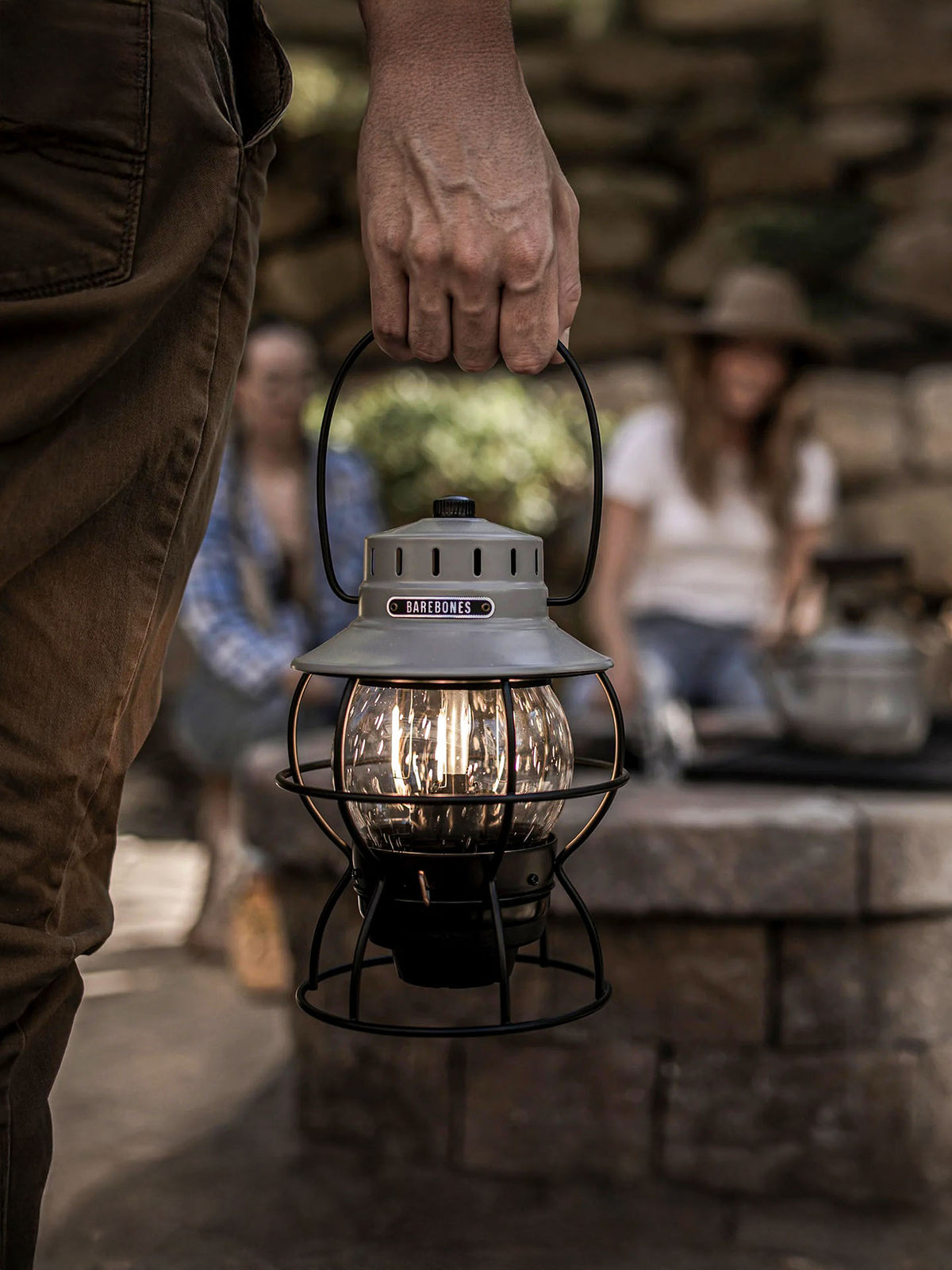 While two people sit near a stone fire pit, a person holds a Barebones Railroad Lantern in Slate Grey outdoors. Its LED bulb casts a warm vintage-style glow.