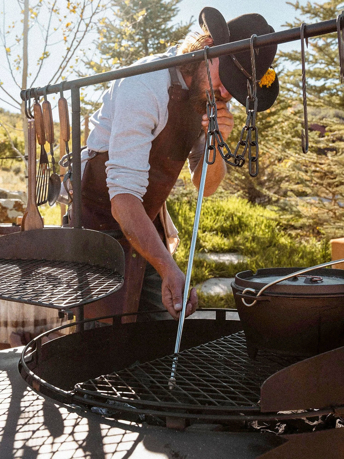 Wearing a hat and apron, a person adjusts a grill over an outdoor fire pit with a pot, grilling utensils, and the Barebones Telescoping Fire Blower nearby.