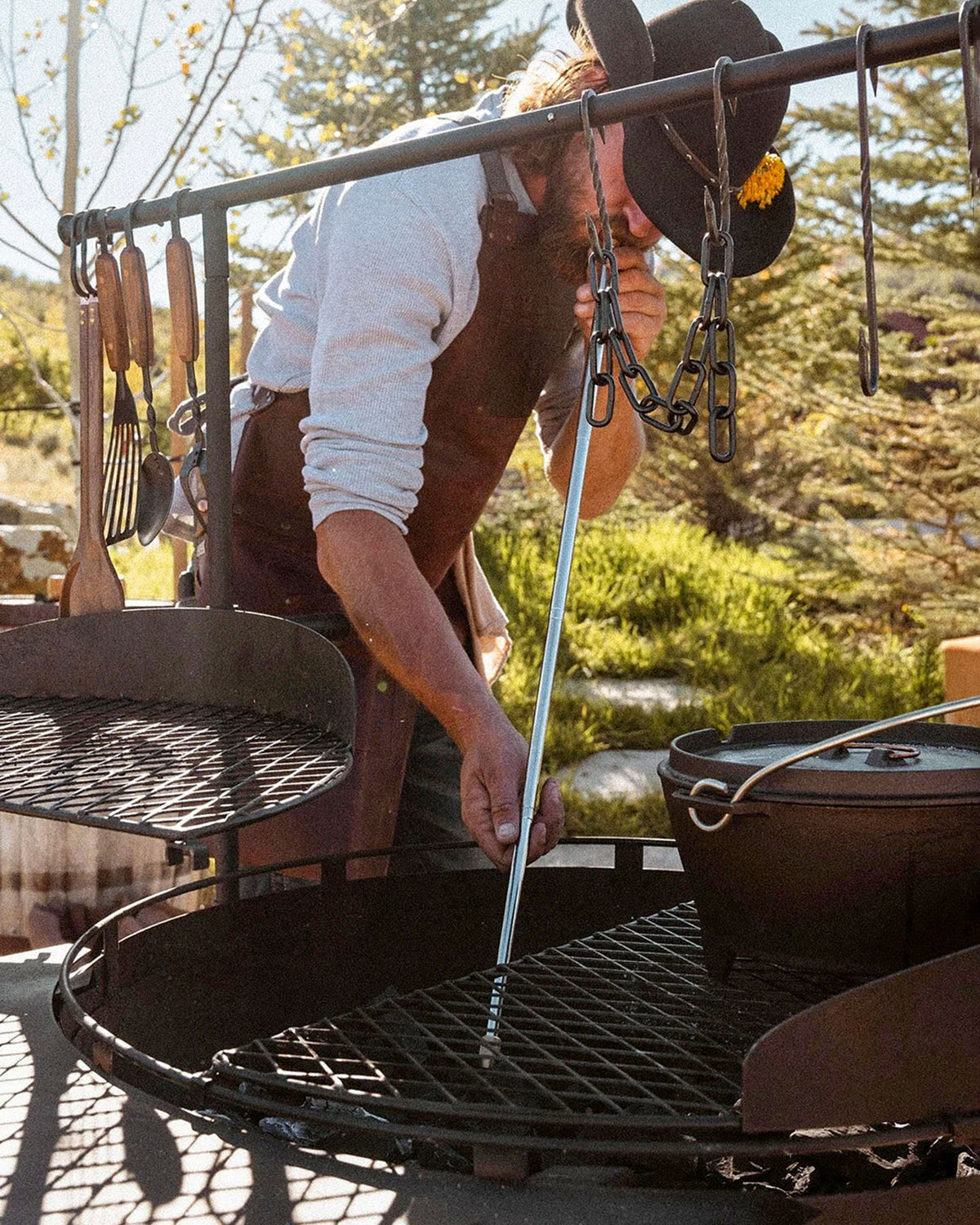 Wearing a hat and apron, a person adjusts a grill over an outdoor fire pit with a pot, grilling utensils, and the Barebones Telescoping Fire Blower nearby.