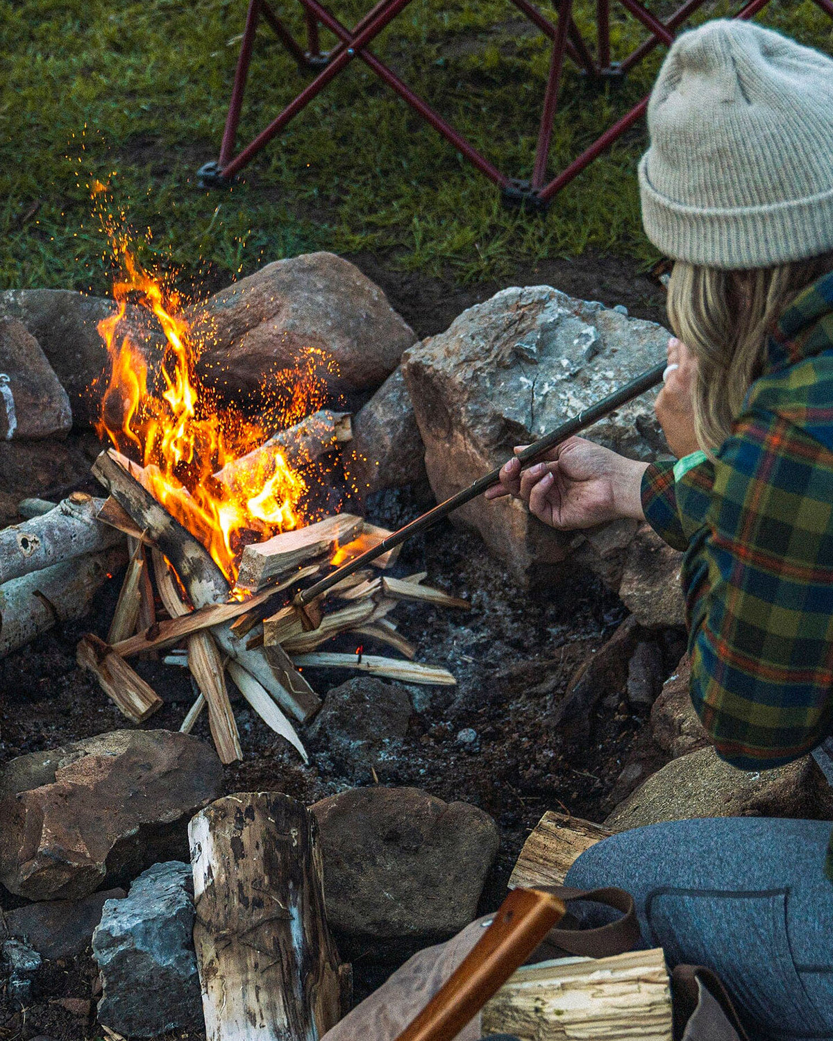Wearing a beanie and plaid shirt, a person uses the Barebones Telescoping Fire Blower to direct air through its pipe, stoking a campfire encircled by rocks and wood.